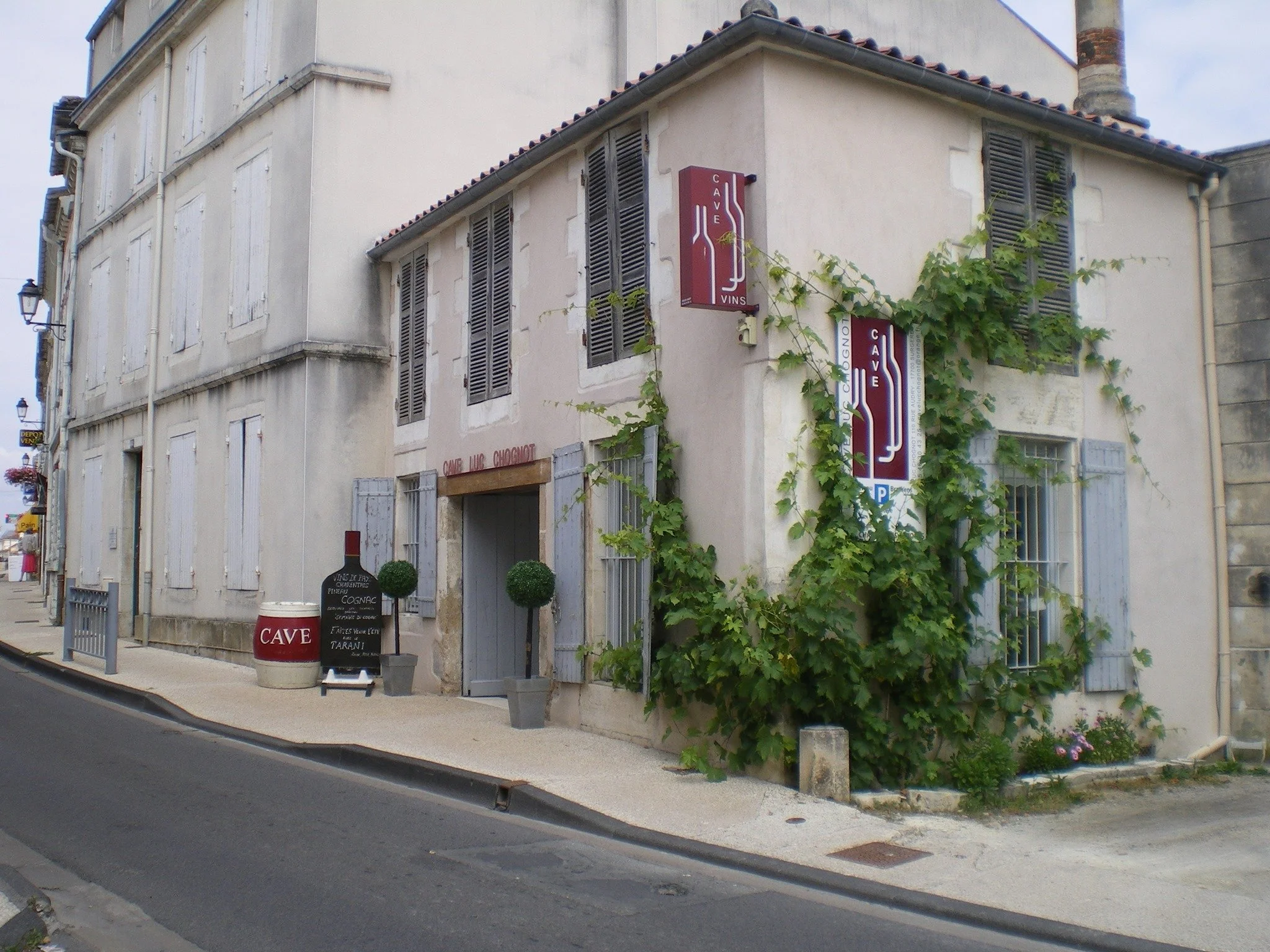 A corner building with a sign reading 'CAVE VINS' and vines growing on the wall, curbside sidewalk, and a street in a quiet European town.