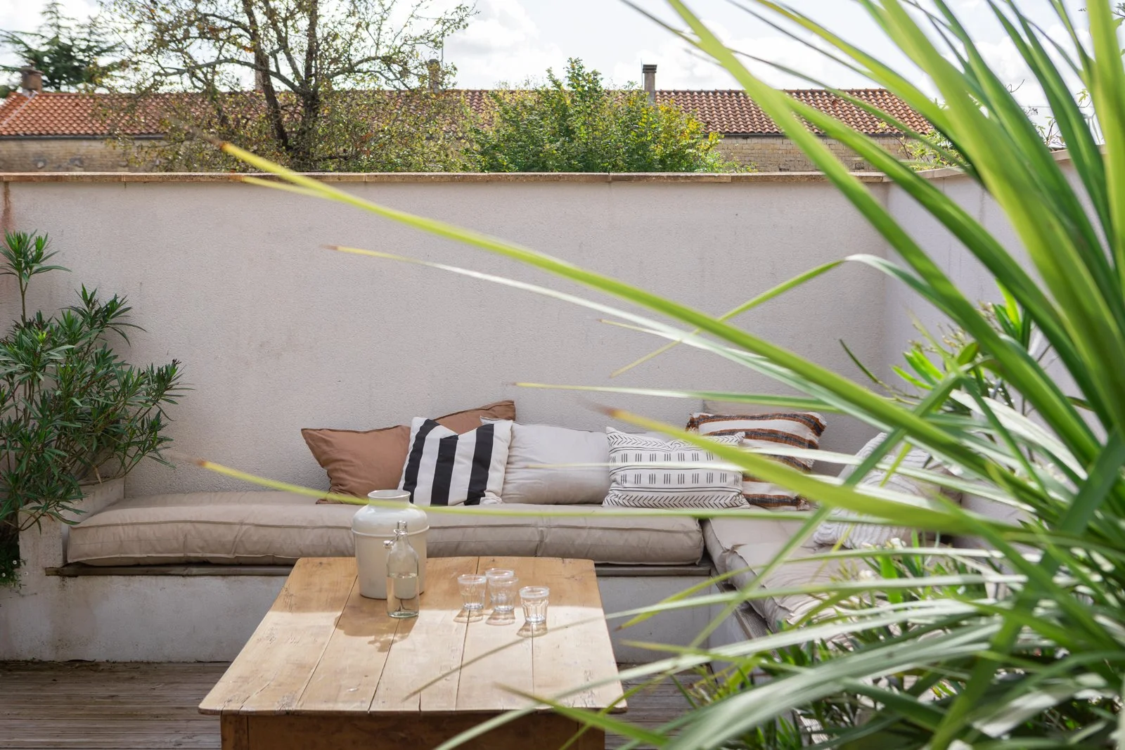 Outdoor patio with a beige couch decorated with patterned and solid throw pillows, a wooden table with glassware and a ceramic jug, surrounded by green plants, and a wall with a red tiled roof in the background.