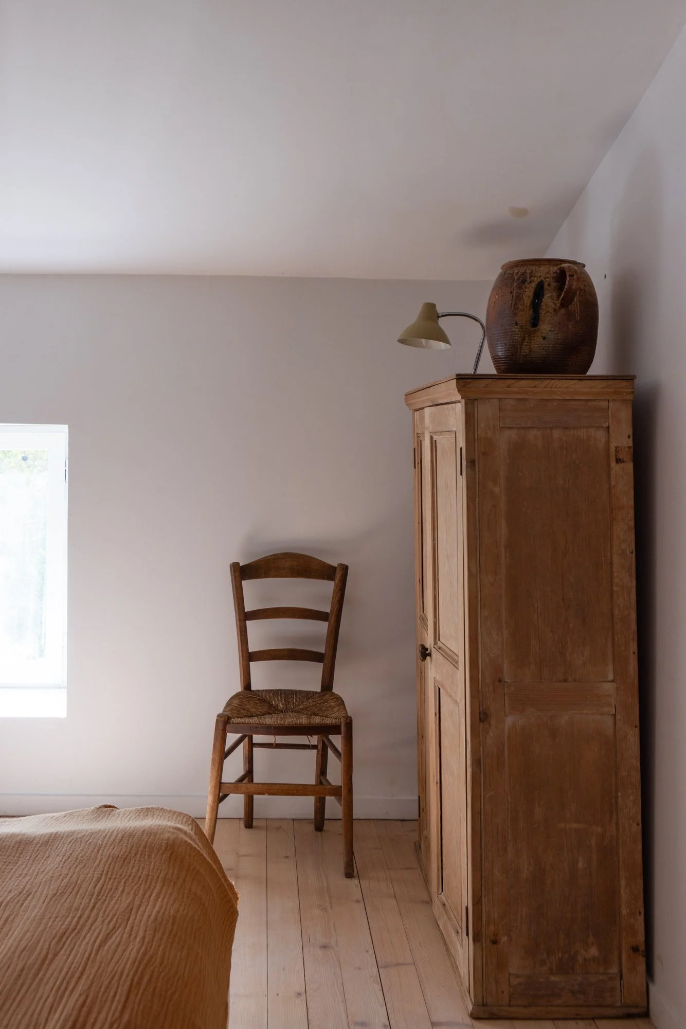 A room with a wooden wardrobe, a wooden chair, a small window, and a large ceramic pot on top of the wardrobe.