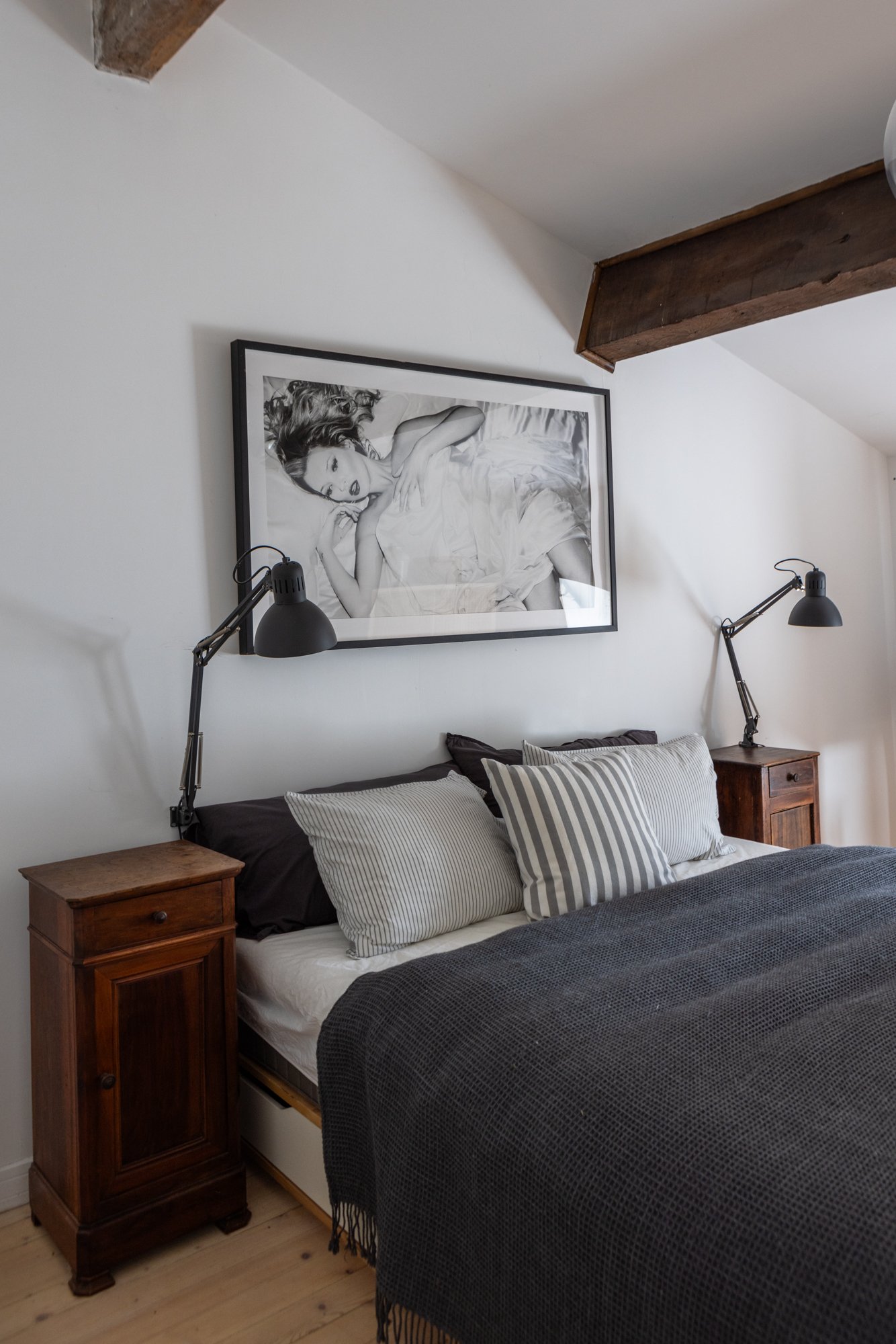 A bedroom with a bed, striped pillows, black blanket, wooden nightstands with black adjustable lamps, and a black and white portrait of a woman on the wall.