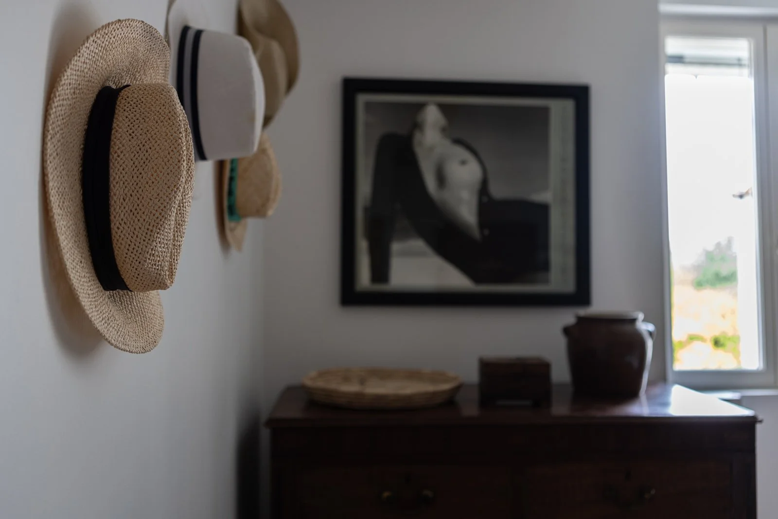 A dark wooden dresser with woven baskets and a large brown ceramic vase on top, a framed black and white photograph of a woman in lingerie hanging above, four straw hats hanging on the white wall to the left, and a window to the right letting in natu