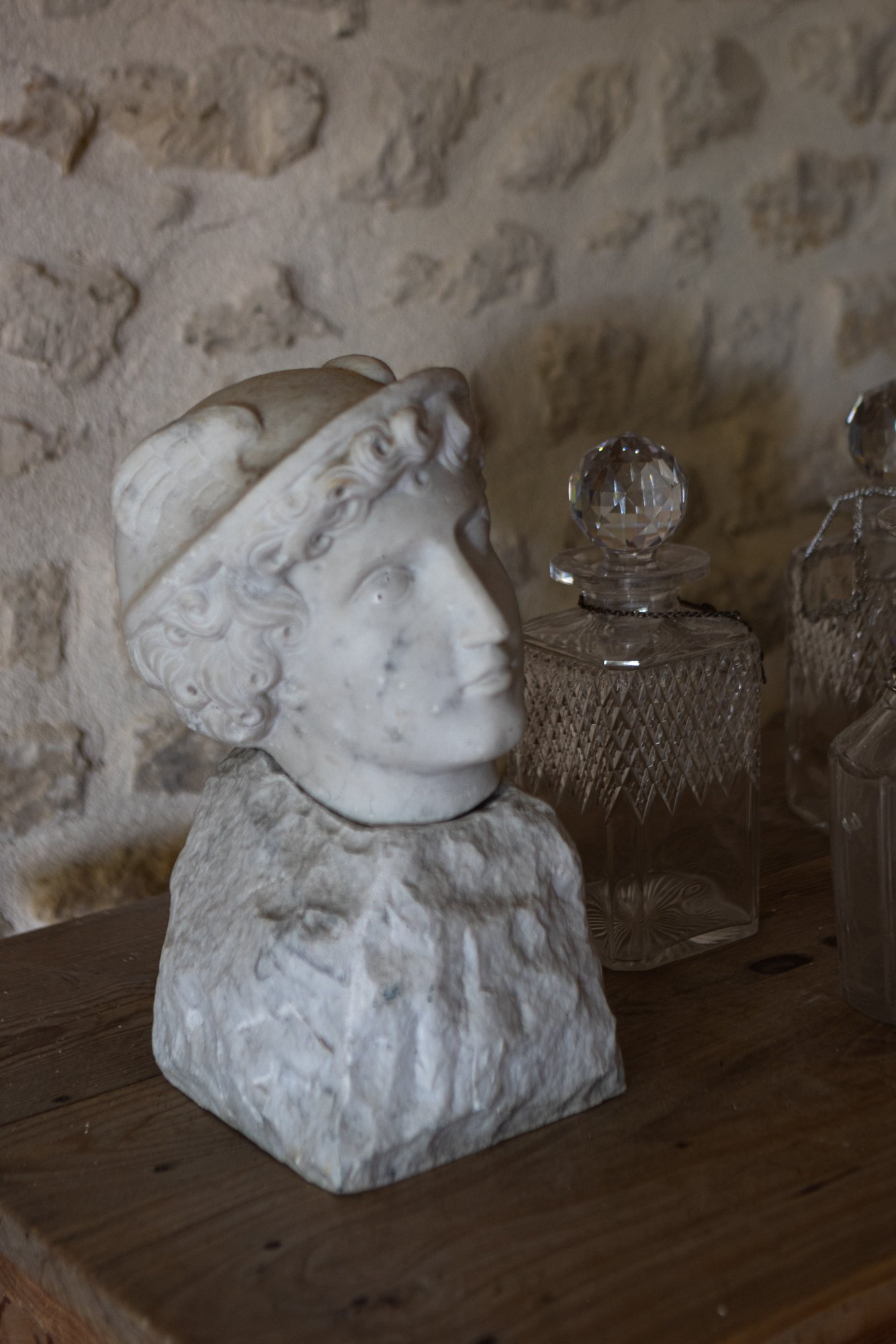 A sculpture of a woman's head with a crown, mounted on a rough stone base, placed on a wooden surface with decorative glass bottles nearby. The background is a stone wall.