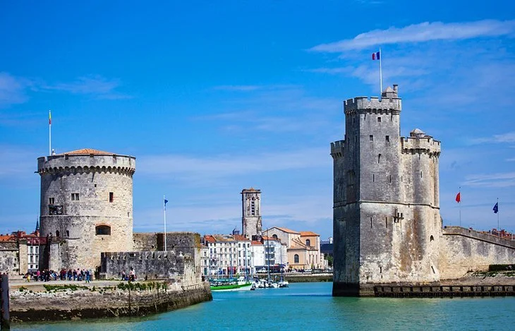 View of historic stone towers and buildings along a waterfront with boats, under a bright blue sky.