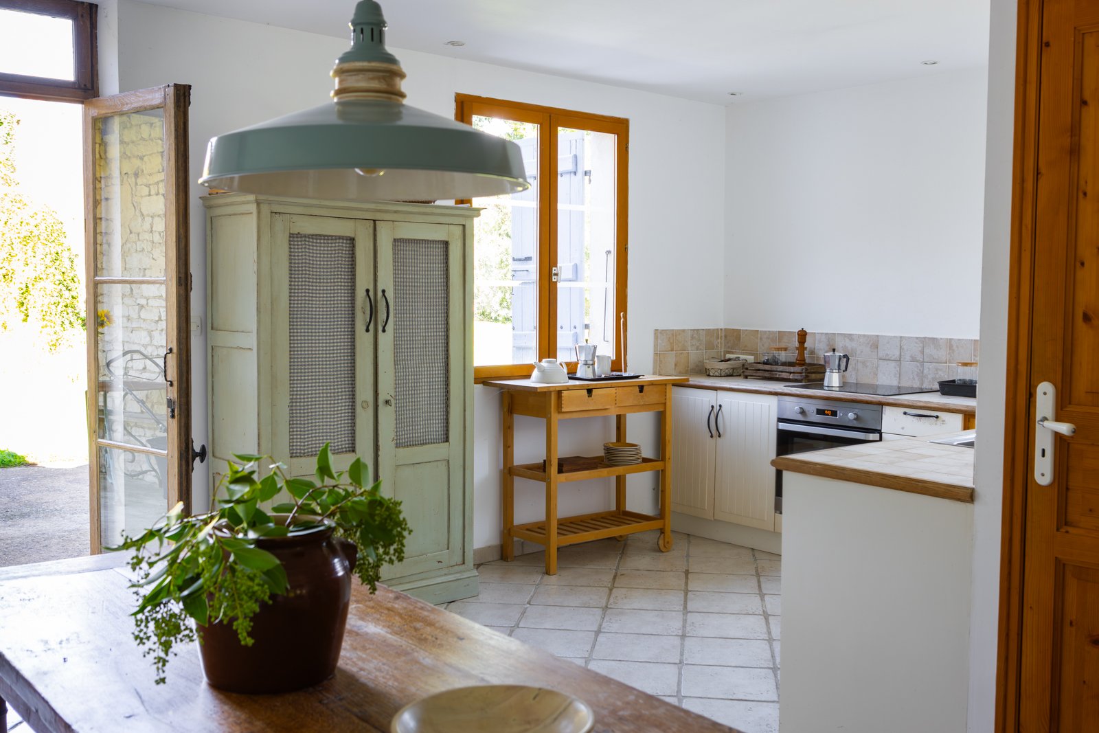 A cozy kitchen with white walls, wooden accents, and sunlight coming through the open window, featuring a white cabinet, a small wooden trolley, and a tiled kitchen counter with kitchen appliances.