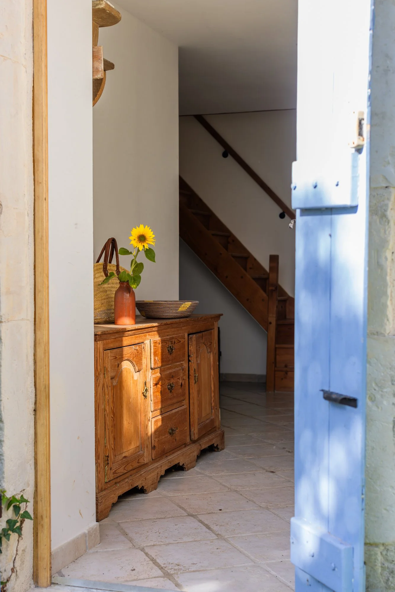 Interior view of a rustic home entrance with a wooden sideboard, a vase of sunflower, and a staircase leading upstairs.