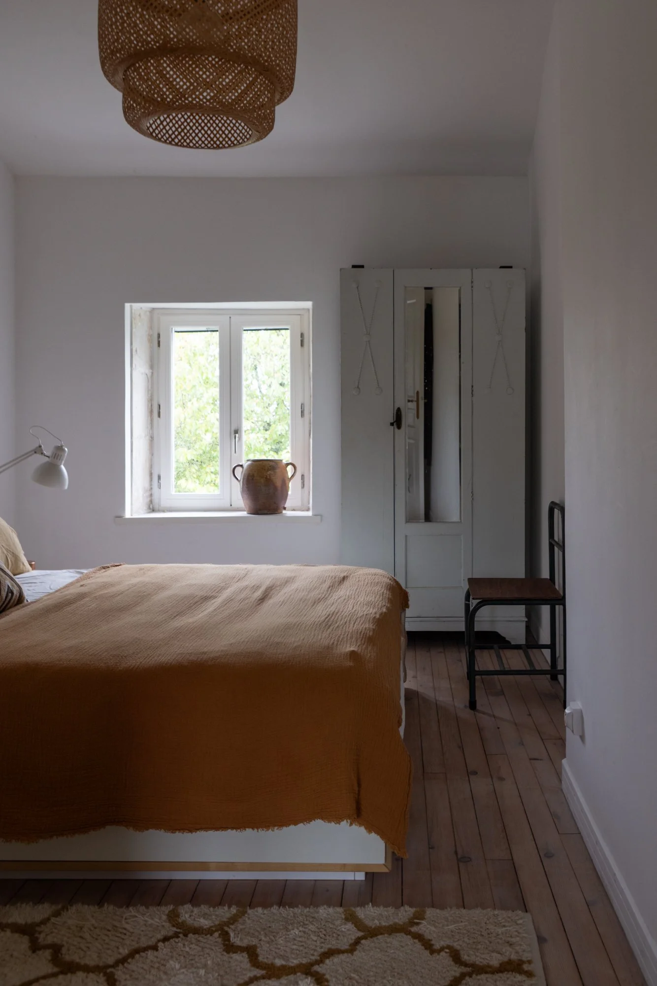 A cozy bedroom with a bed covered in a beige blanket, a window with greenery outside, a ceramic vase on the windowsill, a white wardrobe, a wooden chair, and a woven ceiling light.