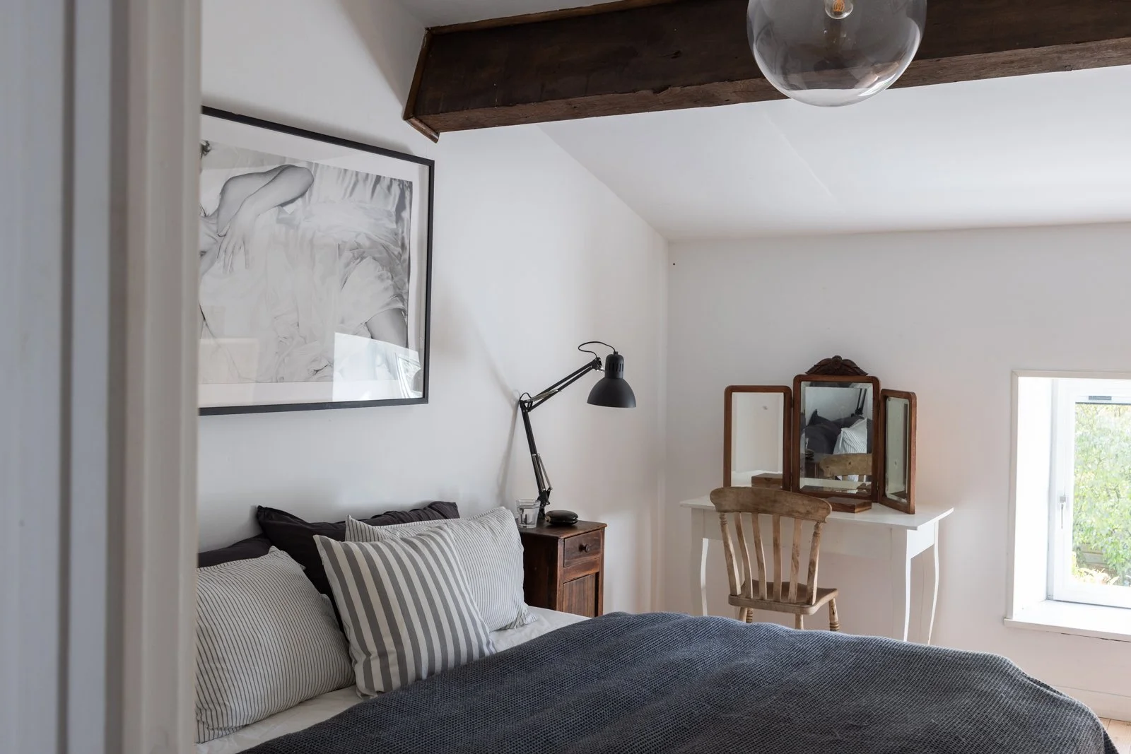 A cozy bedroom with white walls, a bed with striped pillows, a wooden nightstand with a black adjustable lamp, and a small white vanity table with an antique wooden mirror and a wooden chair. There is a window bringing in natural light.