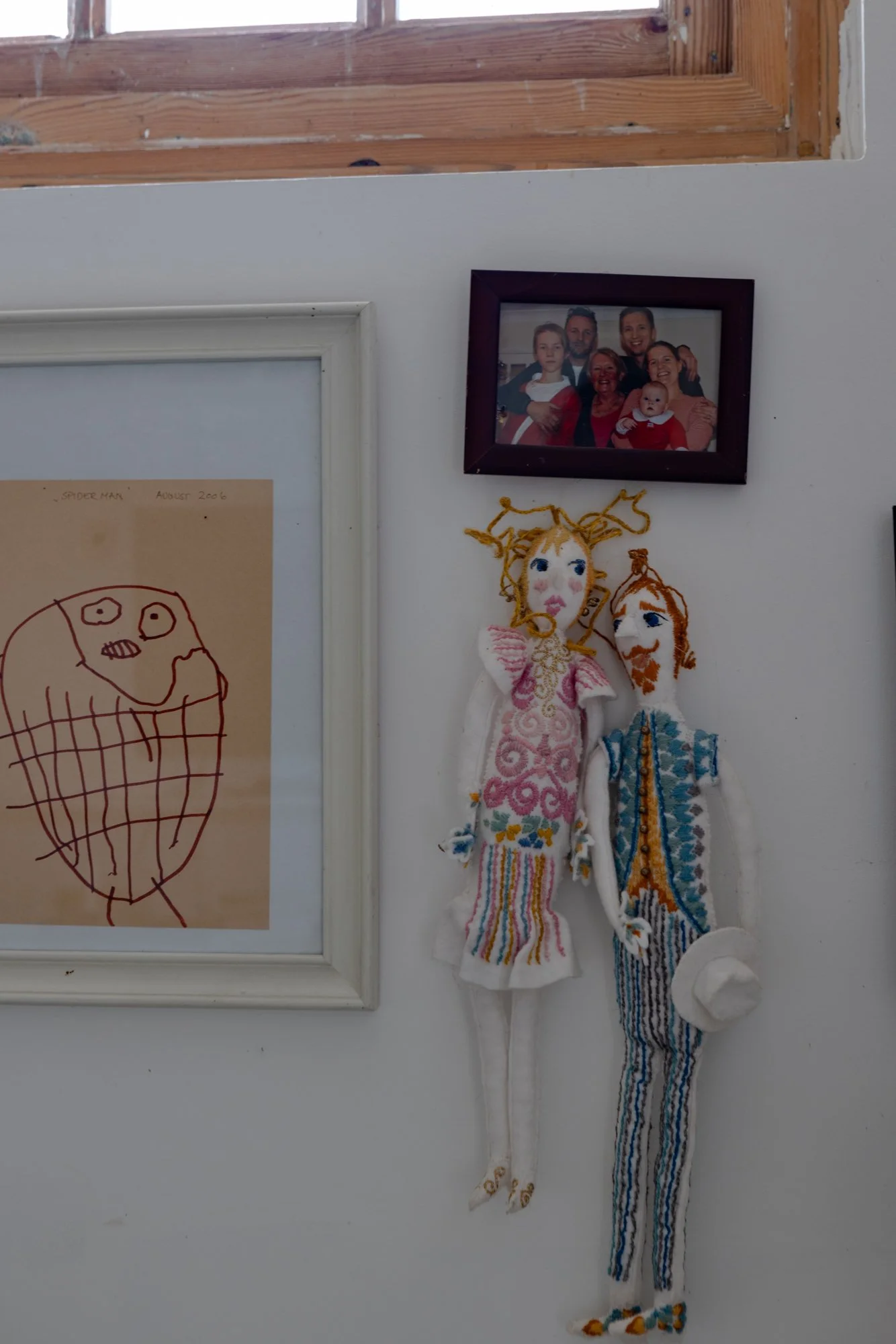 Photo of a wall with a framed family picture, two handmade fabric dolls hanging below, and a child's drawing of Spider-Man on a piece of paper.