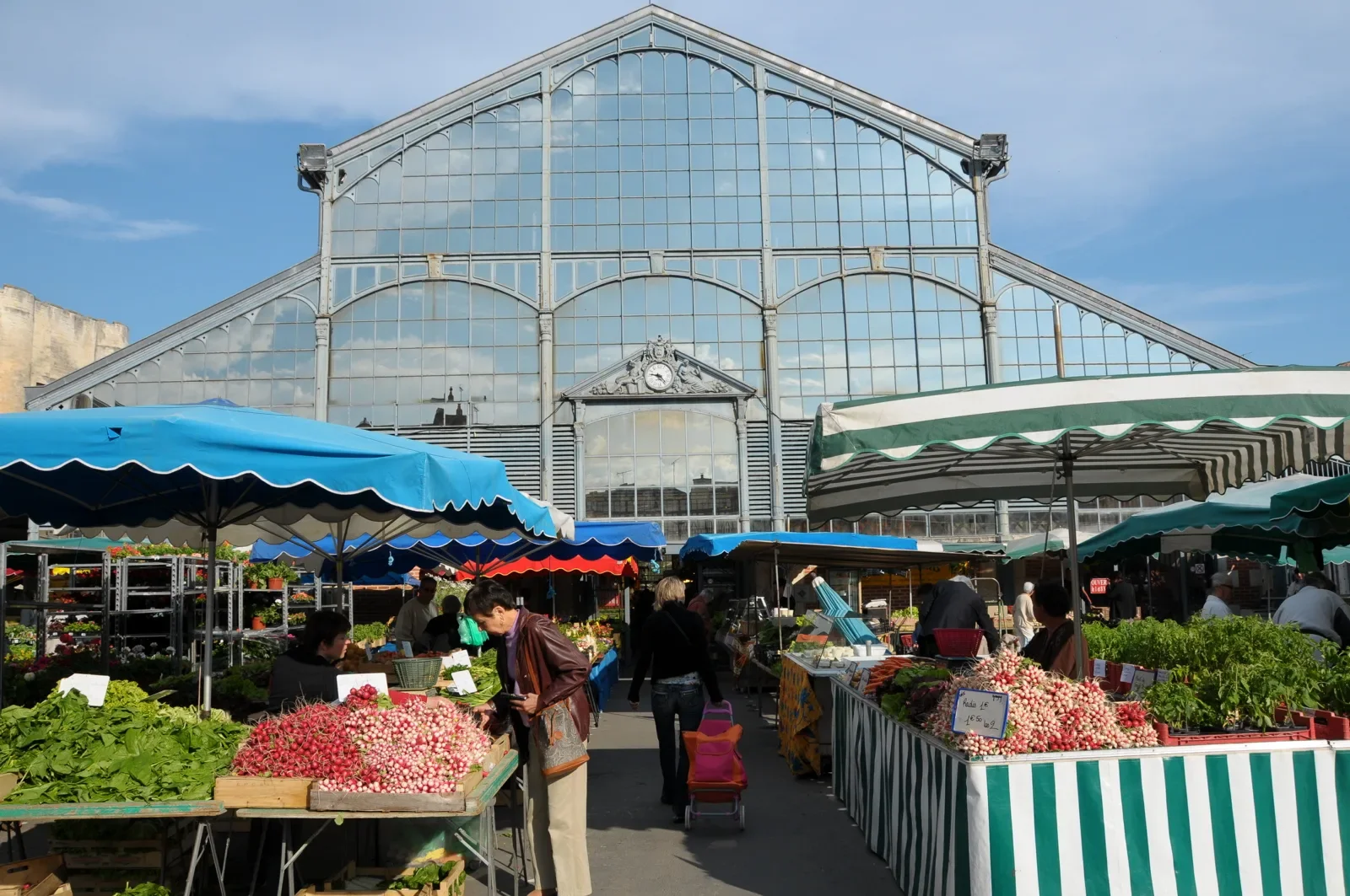 An outdoor farmers market in front of a large glass greenhouse building with blue sky. Market stalls with striped canopies sell vegetables, flowers, and herbs. People browse and shop at the stalls.