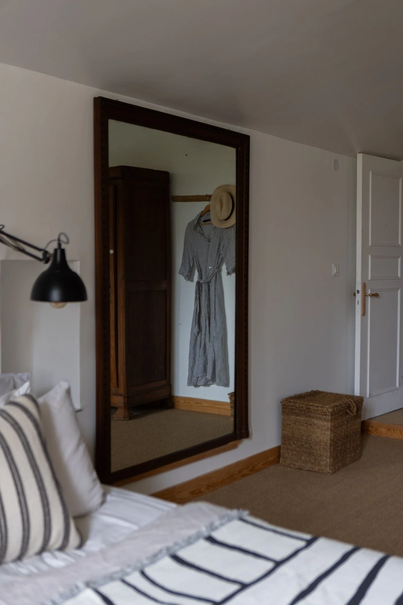 A bedroom with a large wall mirror reflecting a hanging dress and a sun hat, a bed with striped pillows, and a woven storage box on a carpeted floor.