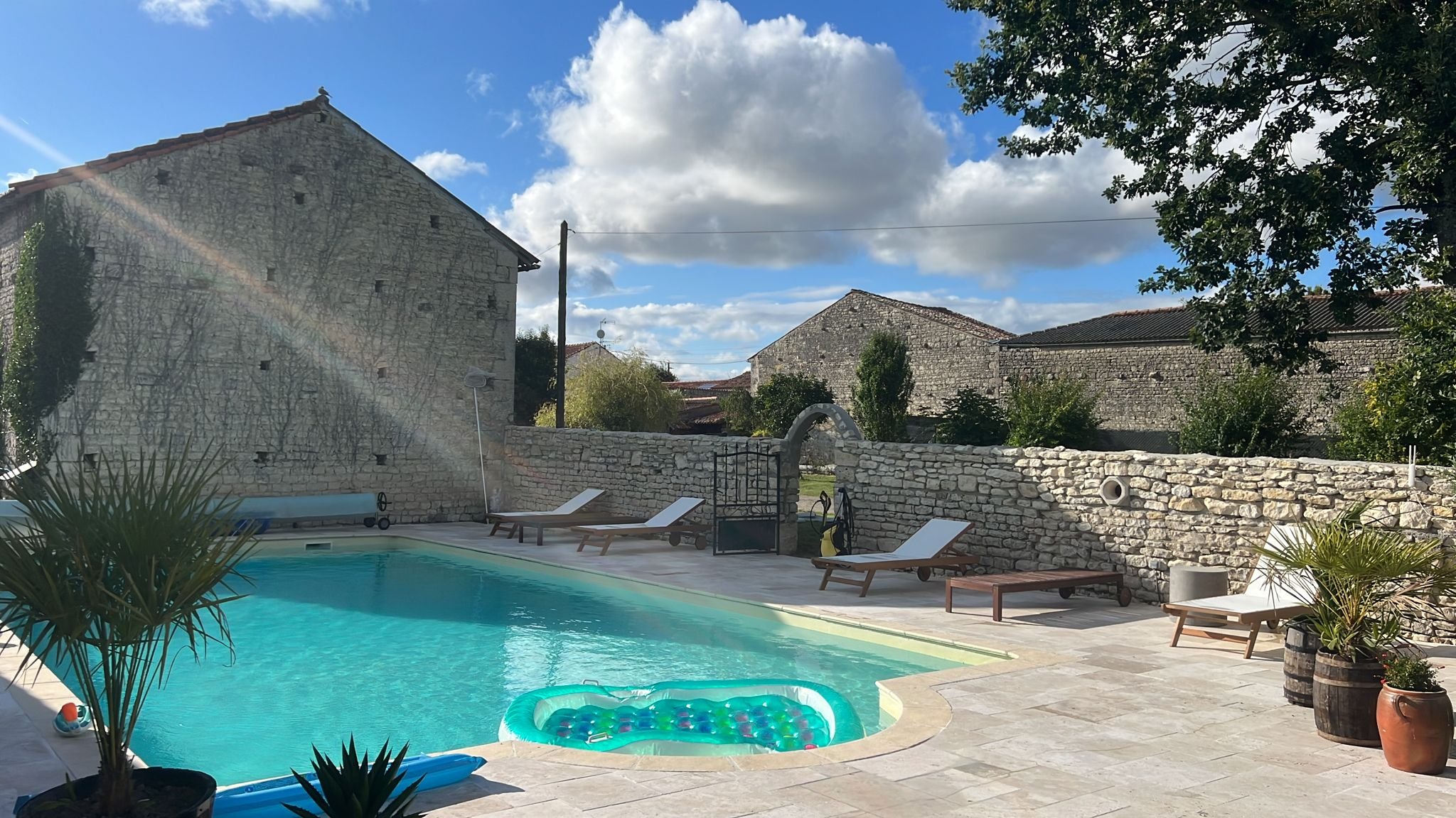 A backyard with a swimming pool, lounge chairs, potted plants, a stone wall, and a blue sky with clouds and a rainbow.