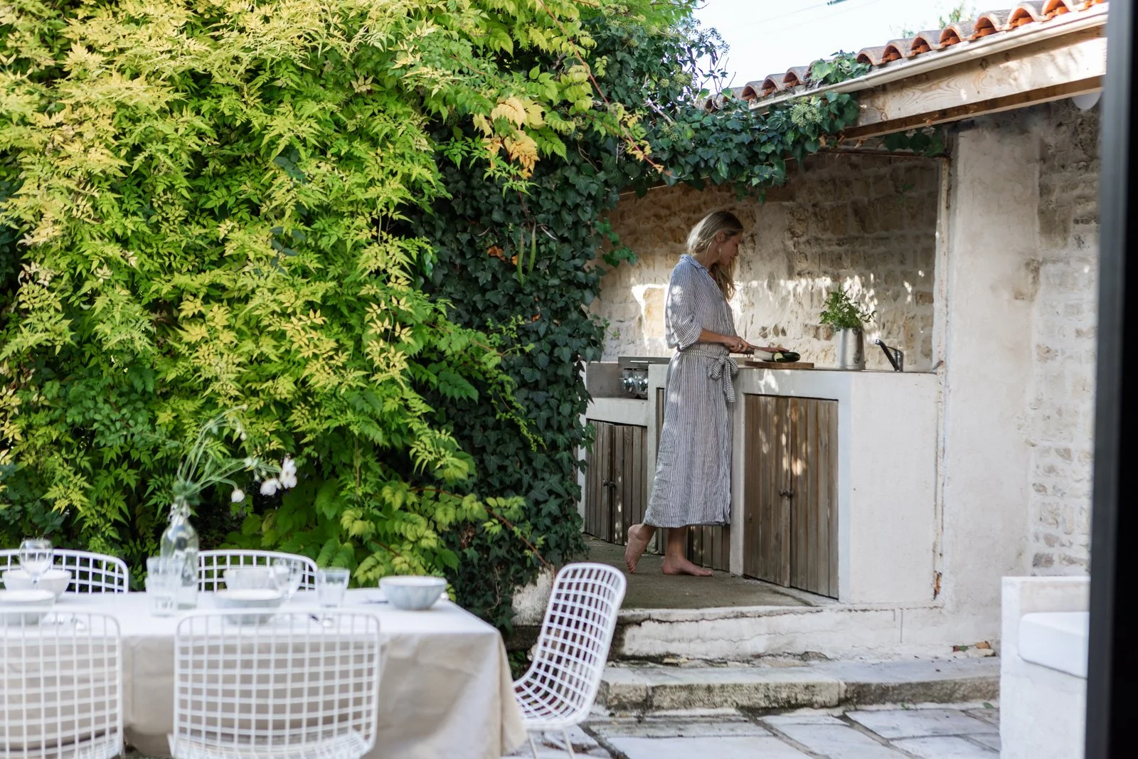 A woman is cooking at an outdoor kitchen area surrounded by greenery, with a dining table in the foreground.