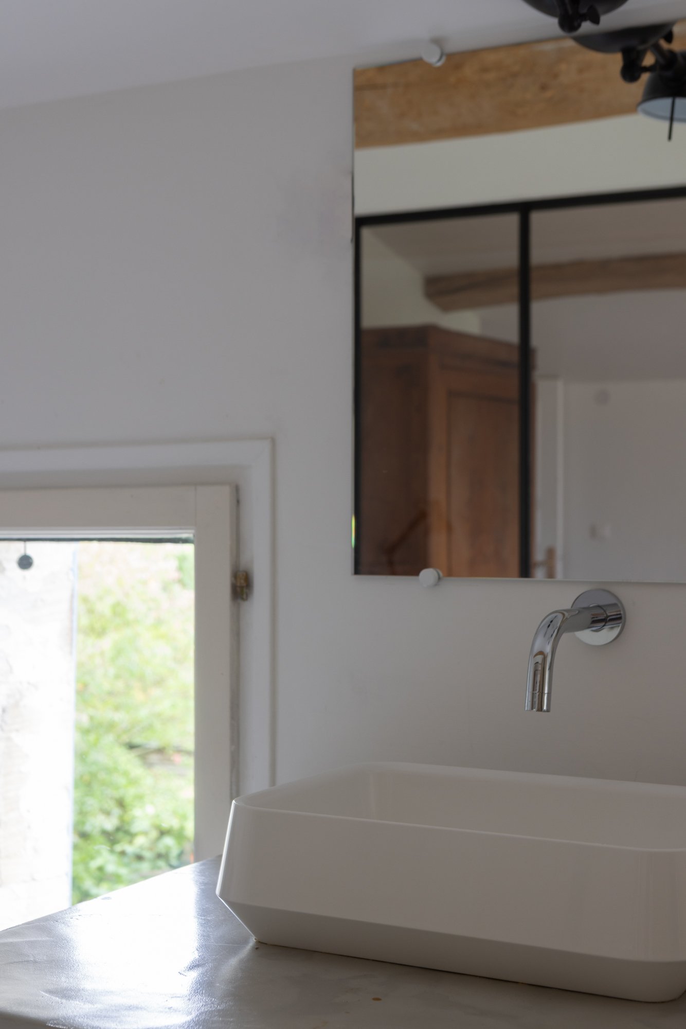 A modern white rectangular sink with a curved interior, mounted on a white wall in a minimalist bathroom, with a chrome faucet above it. A mirror reflects wooden elements and a ceiling beam.