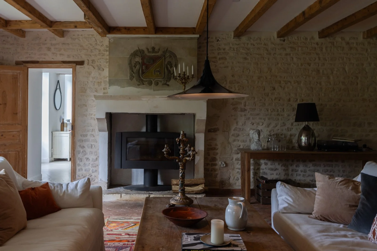 Living room with stone walls, wooden beams, a fireplace with a black stove, a chandelier, a table with decorative items, and sofas with cushions, styled in vintage decor.