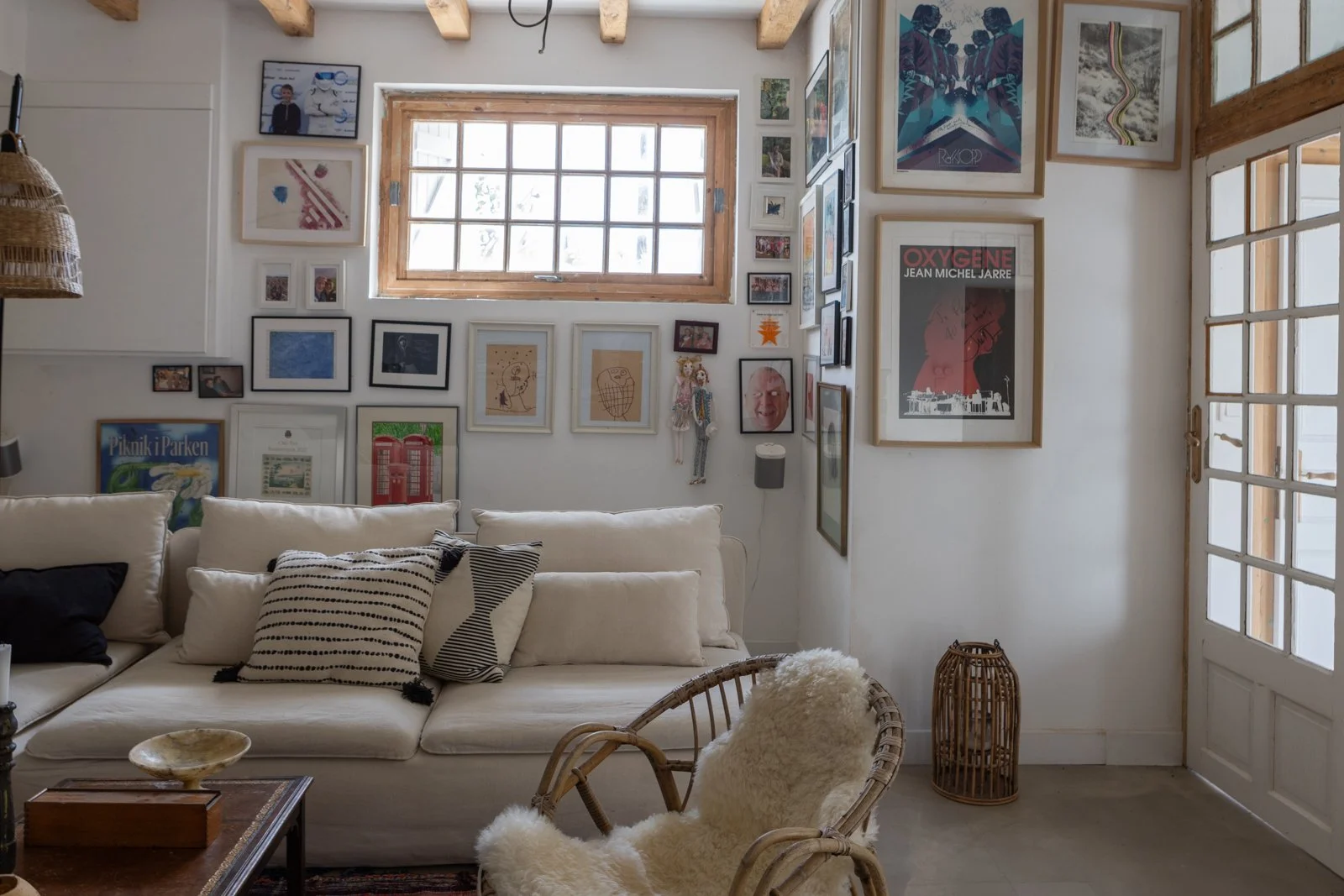 Cozy living room with beige sofa, black and white striped pillows, rustic wooden coffee table, rattan armchair with sheepskin cushion, gallery wall with framed art and photos, window with wooden trim, door with glass panes, and wicker basket decor.