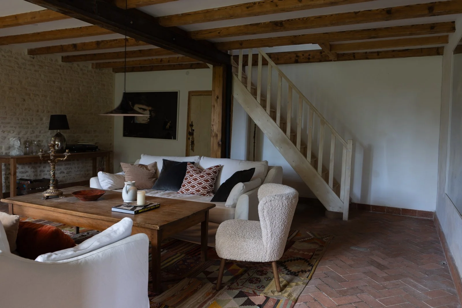 Living room with white sofa, black and patterned pillows, wooden coffee table, white armchair, staircase with wooden railing, exposed brick wall, and modern artwork.