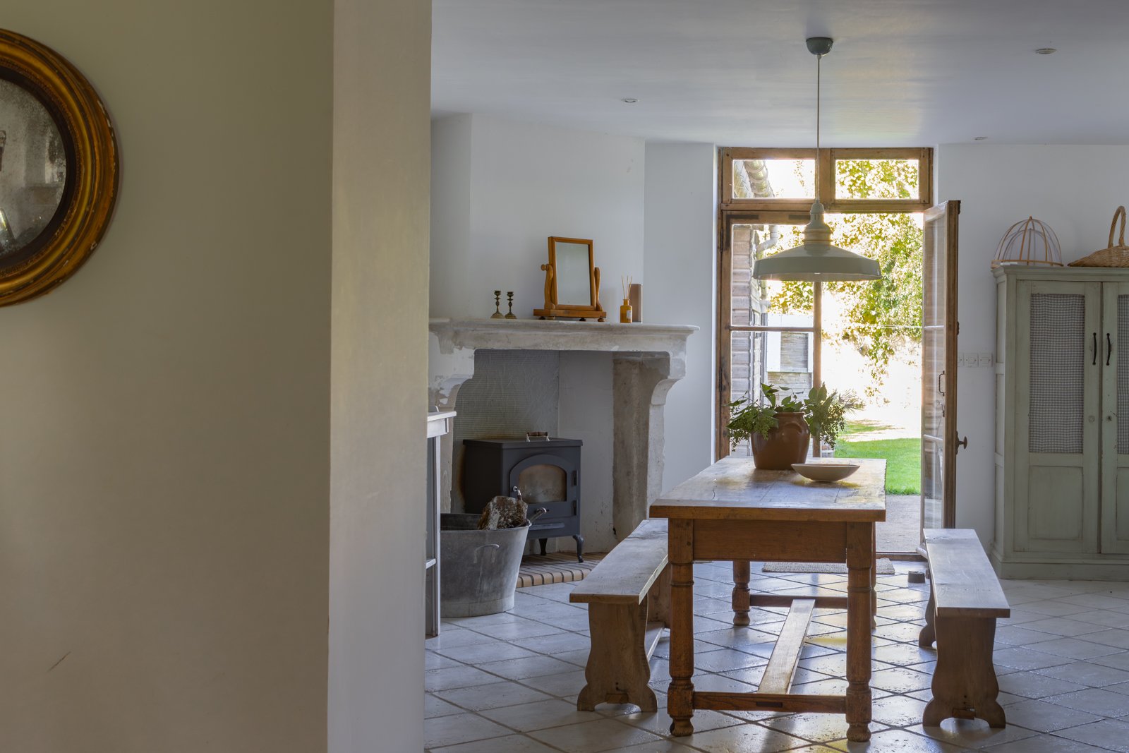 A cozy, rustic kitchen with a wooden dining table, benches, and a stone fireplace. Natural sunlight comes through open wooden doors leading outside.