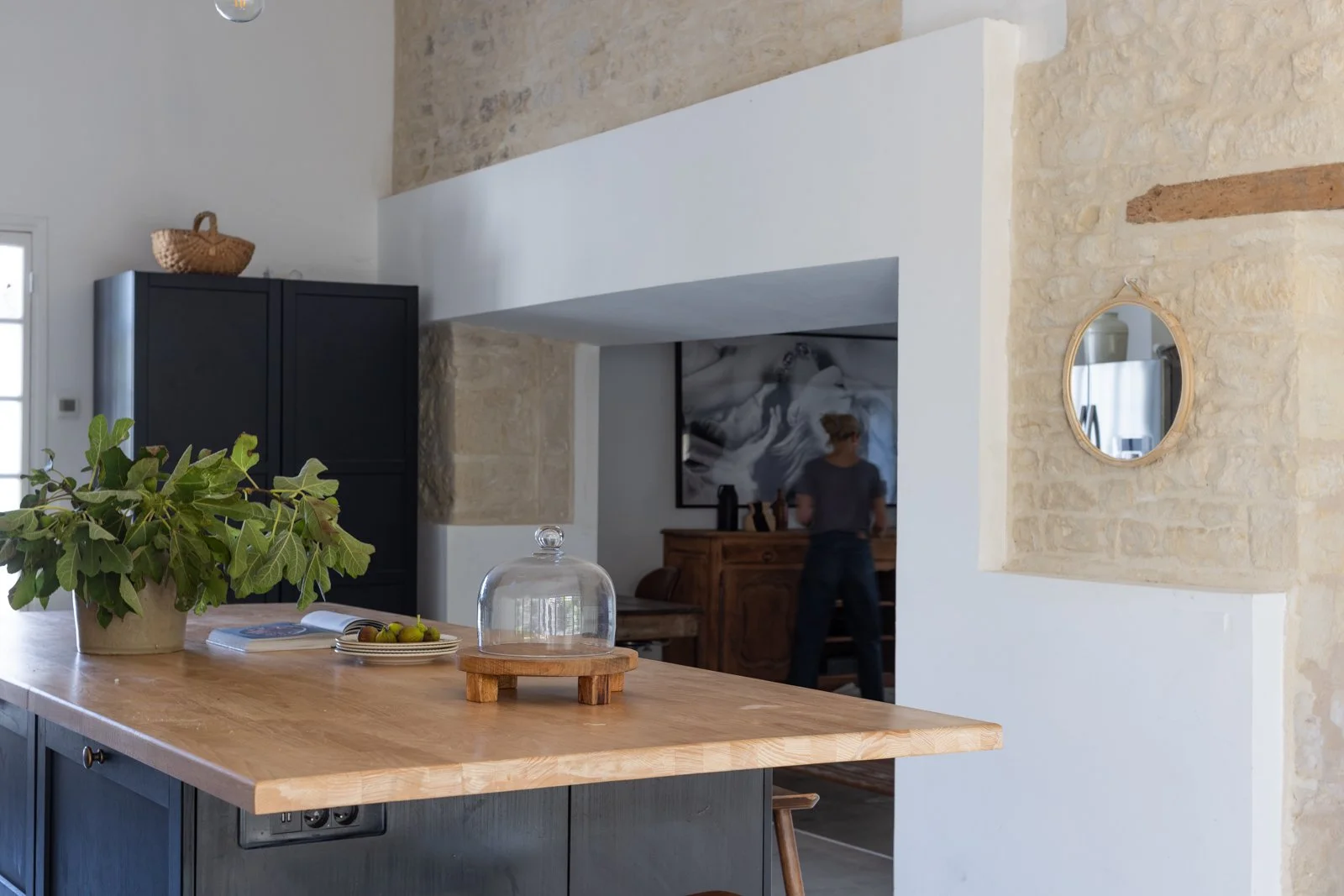 A kitchen with a wooden island, a potted plant, a glass cloche, and a fruit plate. In the background, a person is standing near a wooden cabinet and an abstract painting.