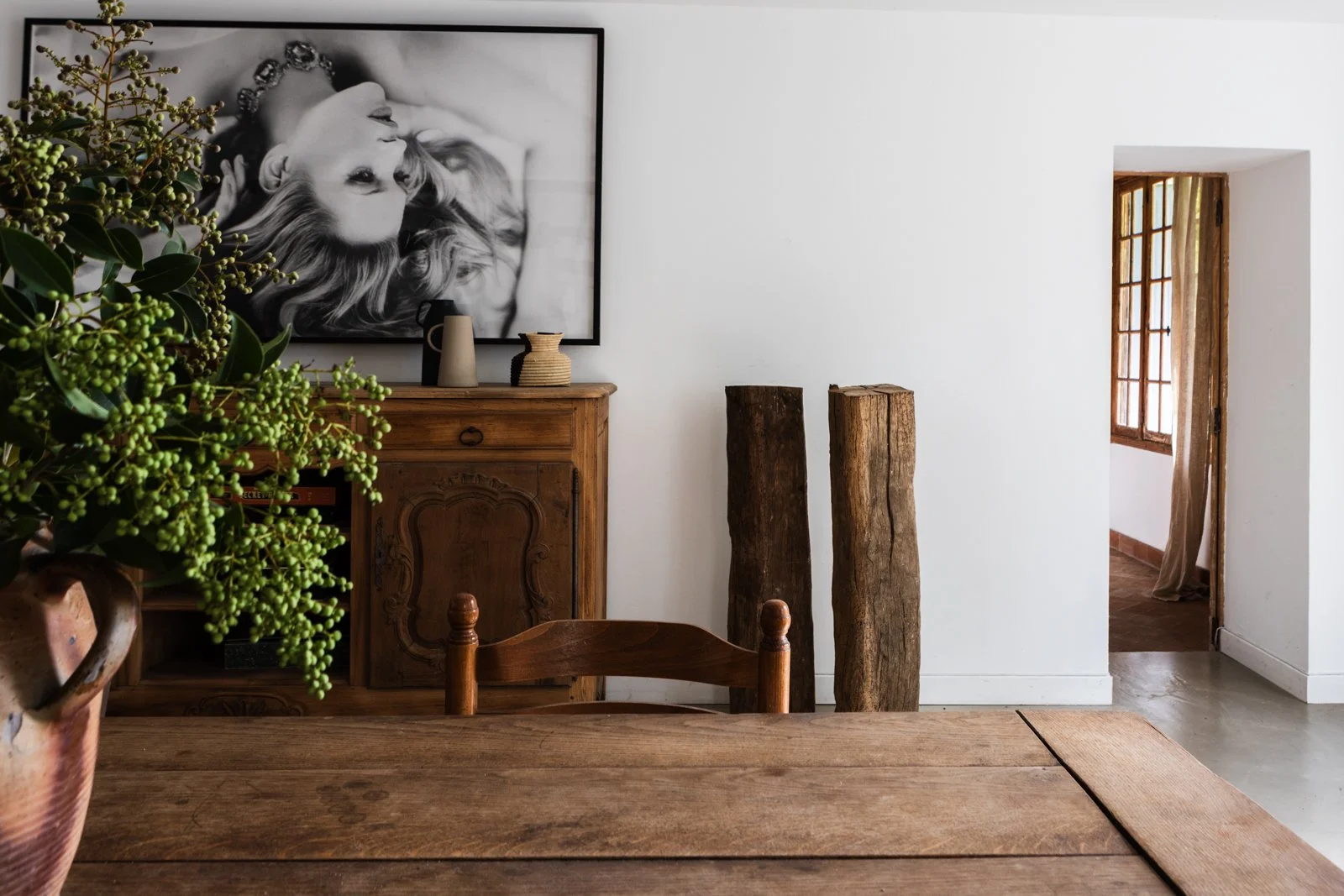 Interior of a room with a wooden table, a wooden sideboard decorated with pottery, a large black-and-white portrait of a woman on the white wall, and two large tree logs standing against the wall. There is a window with multiple panes and a doorway w