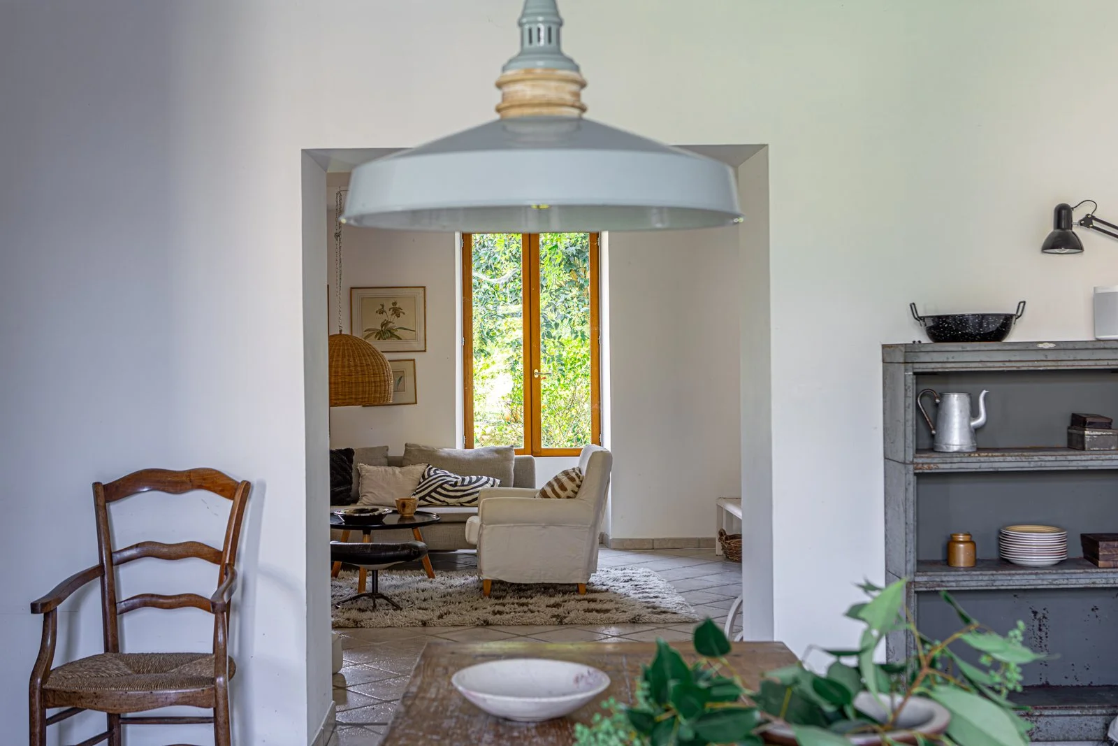 View through a doorway into a cozy living room with a large window, sofa, armchair, and decorative pillows, with a wooden table and a hanging pendant lamp in the foreground.