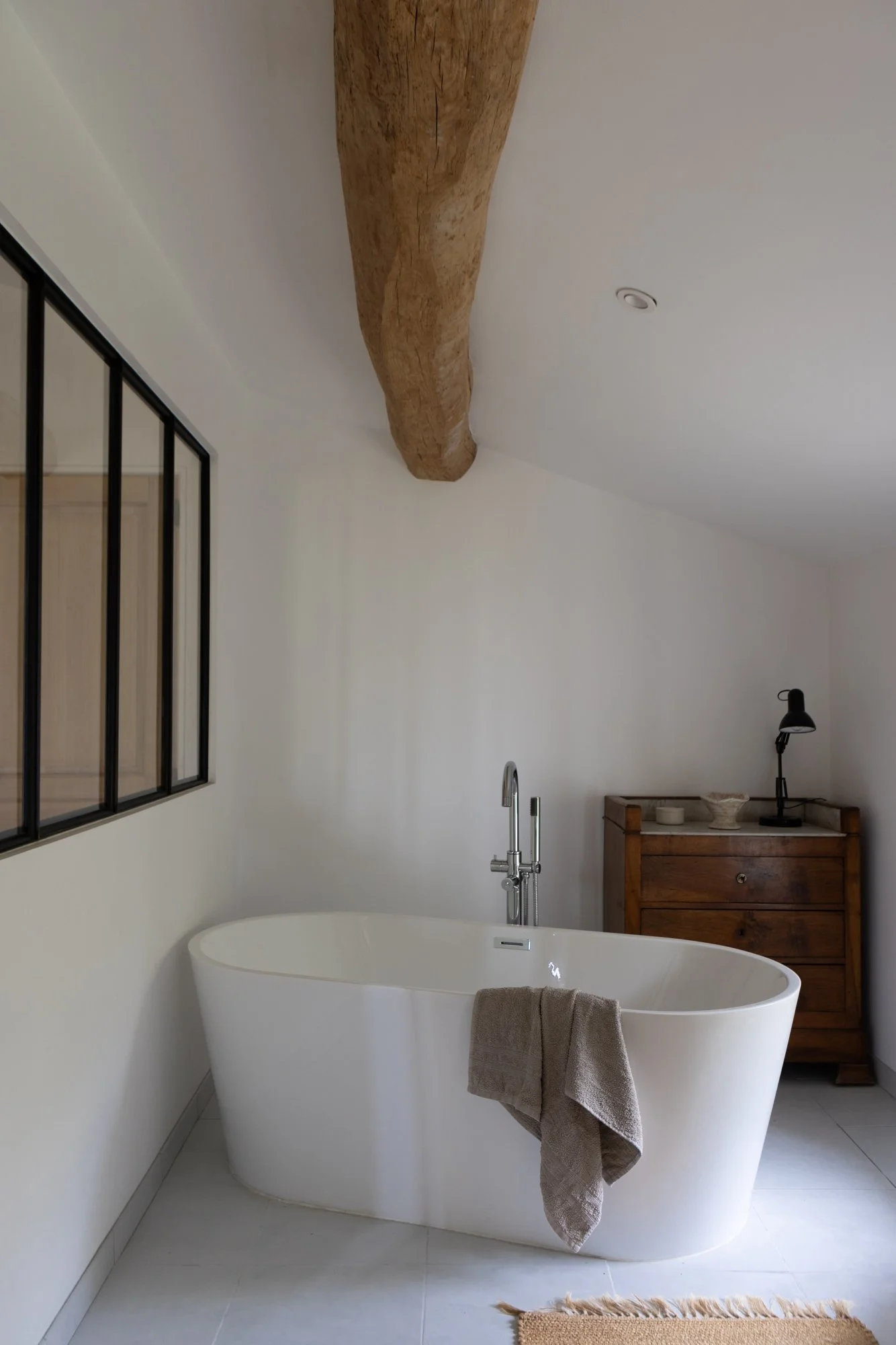 Modern bathroom with a freestanding bathtub, a small wooden cabinet, a black desk lamp, and a large wooden beam on the ceiling.