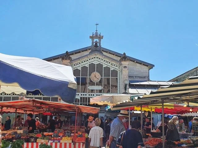 Farmers' market stalls with people shopping, market tents, and a historic building with a clock in the background.