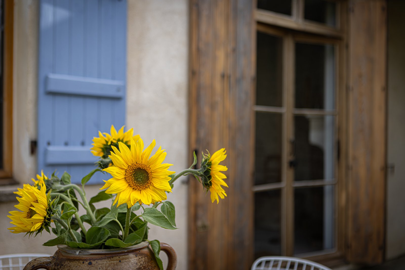 Sunflower bouquet in a rustic clay pot on a table in front of a weathered building with a partially open wooden window and a blue door.