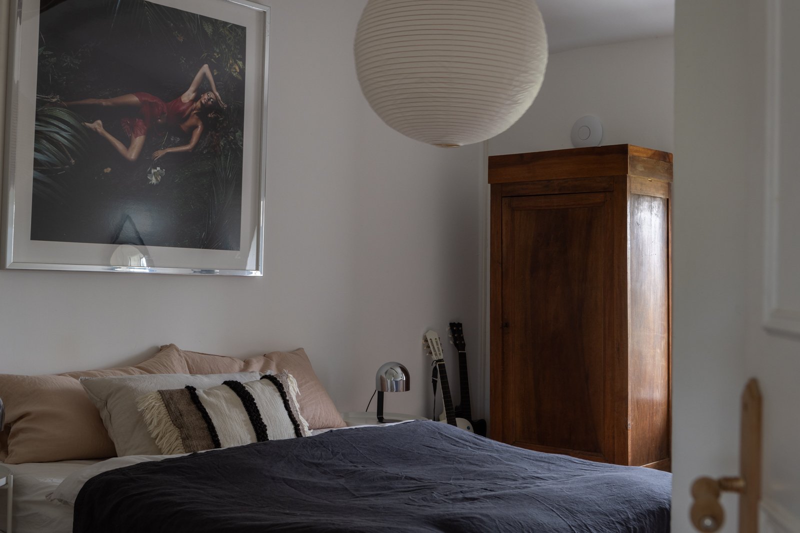 A bedroom with a framed photo of a woman in a red dress hanging above the bed, a white paper lantern ceiling light, a wooden wardrobe, two guitars, a modern desk lamp, and pillows on the bed with black and white striped pillow, beige pillows, and a d