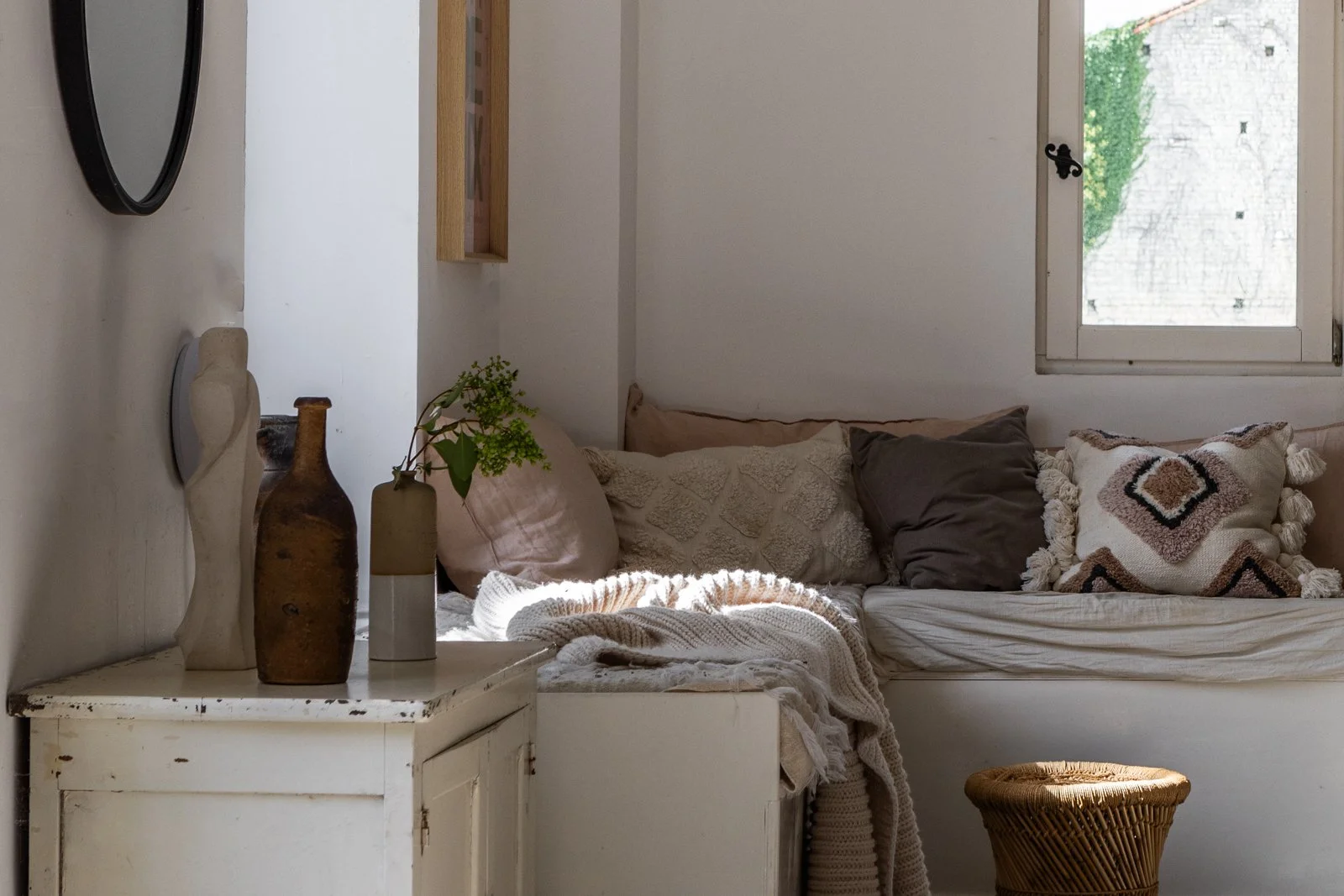 Sunlit corner in a cozy living space with a white wooden sideboard holding decorative vases and a green plant. A beige sofa with assorted cushions and a textured throw blanket is set against the wall, with a small wicker stool in front and a window l