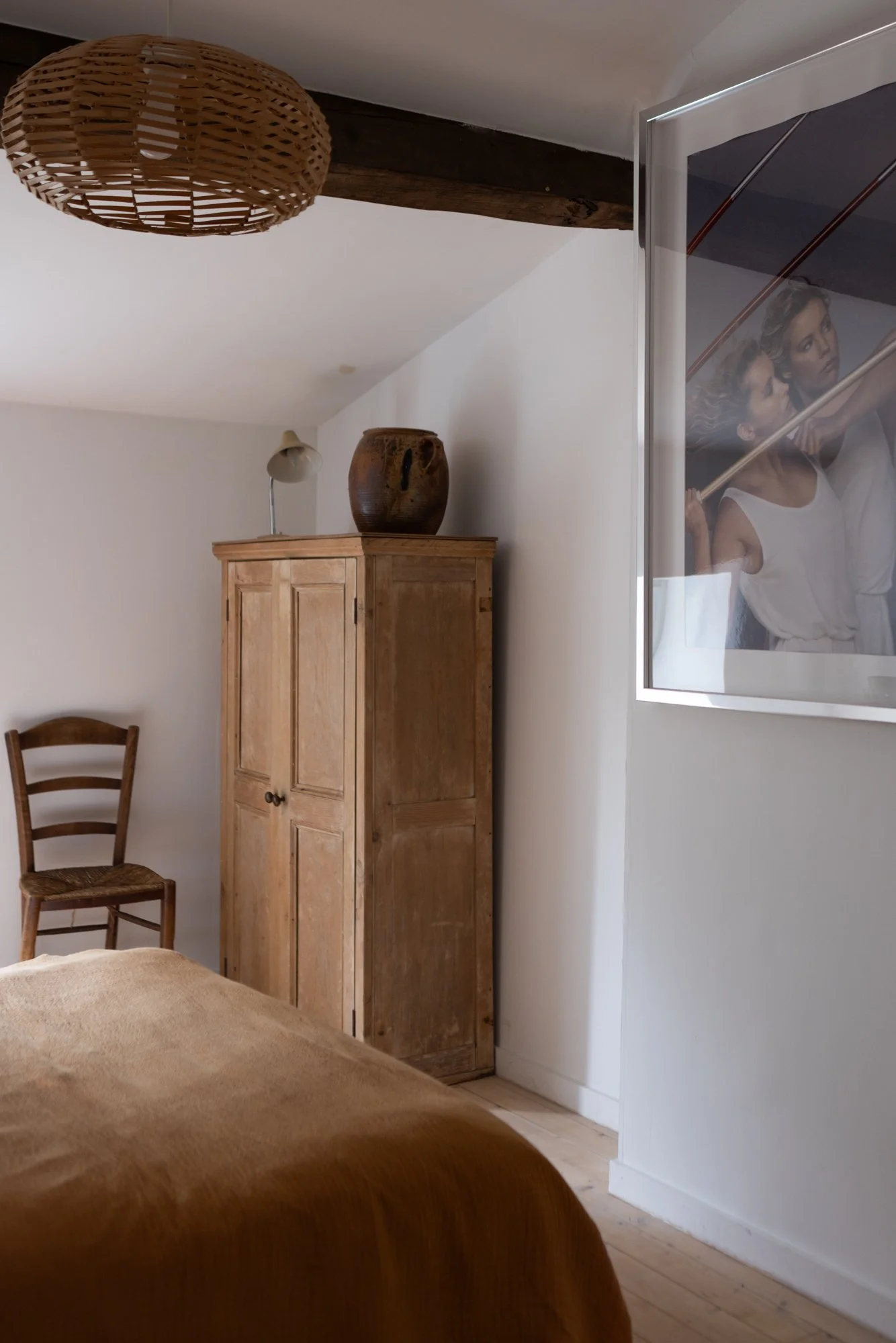 A cozy bedroom with a wooden wardrobe, a chair in the corner, a bed covered with a brown blanket, a framed photograph on the wall, and a woven ceiling light.
