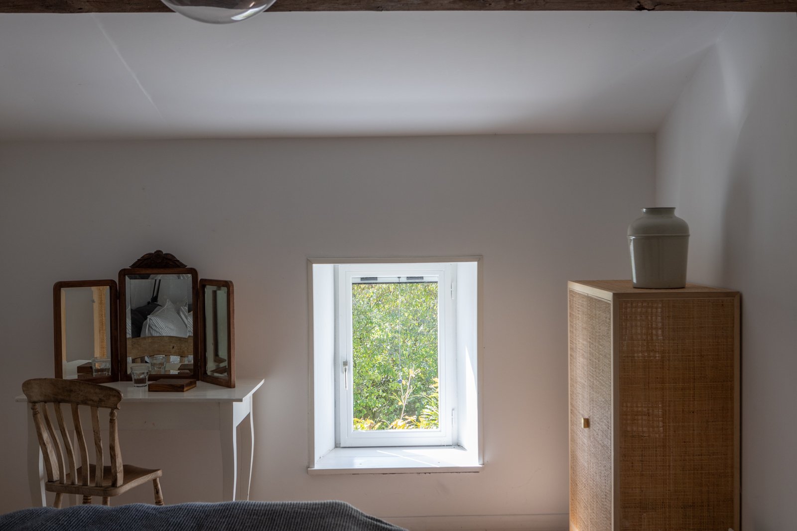 A bedroom with white walls, a small window showing green trees outside, a wooden chair, a white dressing table with a three-panel mirror, a wooden cabinet with a white vase on top, and part of a bed in the foreground.