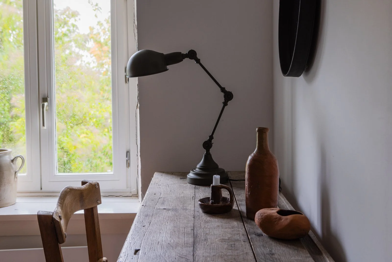 A wooden table near a window with a view of greenery outside, decorated with several pottery vases and a black adjustable desk lamp.