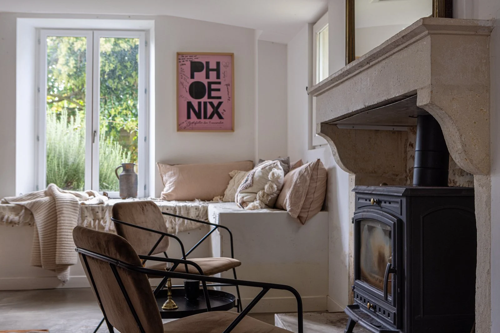 Cozy living room with large window, beige and cream cushions, a pink Phœnix poster on the wall, a fireplace, and two brown chairs.