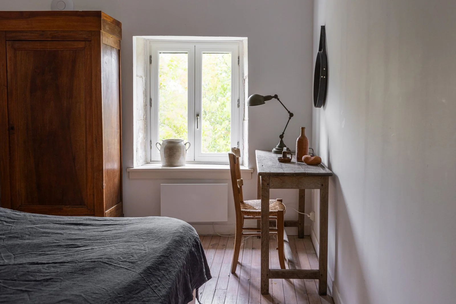 A cozy bedroom corner with a wooden wardrobe, a small desk with a vintage lamp, pottery, and a chair. A window with greenery outside lets in natural light.