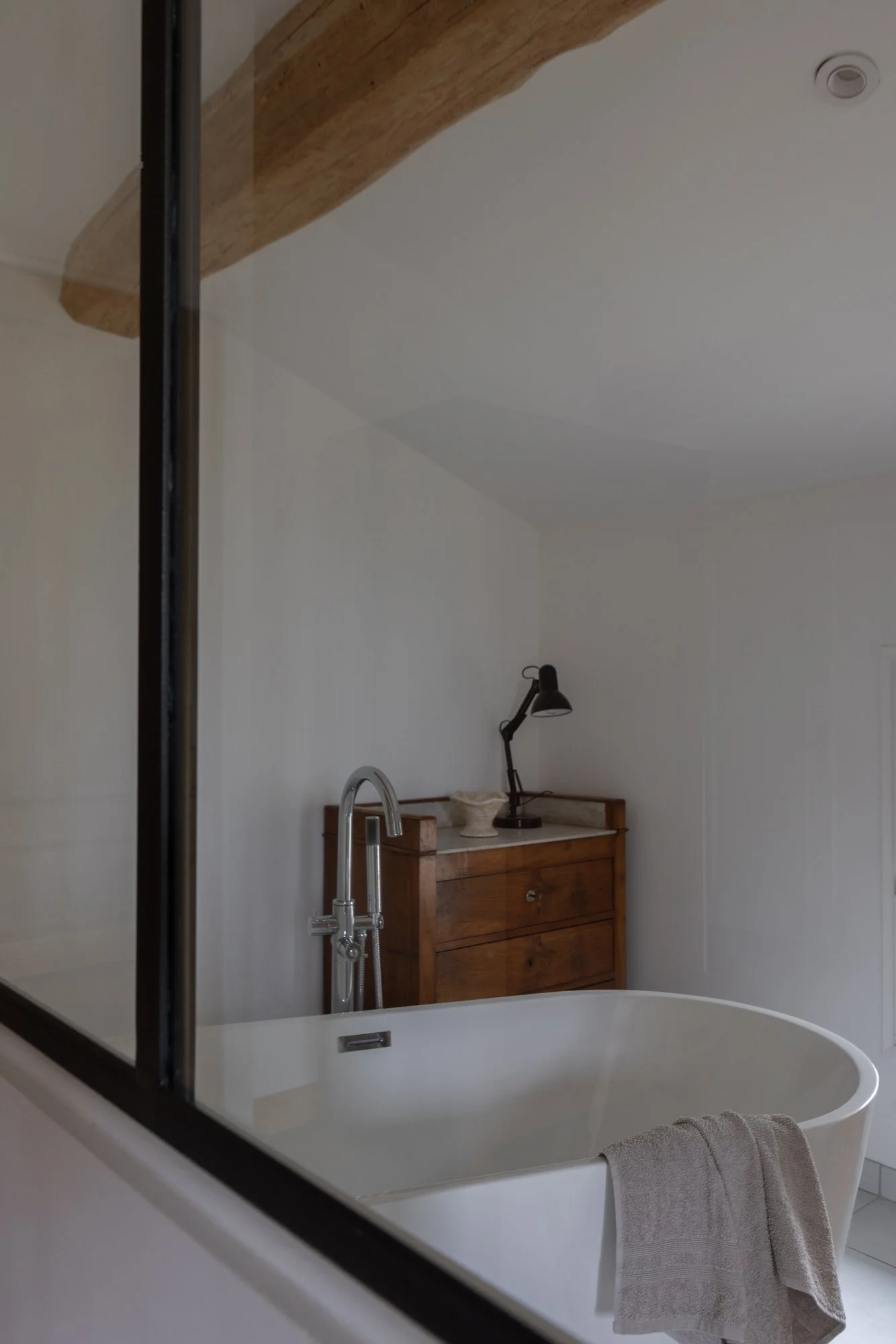 A bathroom with a free-standing bathtub, a towel hanging over its edge, a wooden dresser with a black desk lamp, a white bowl, and a decorative item on top, viewed through a glass partition.