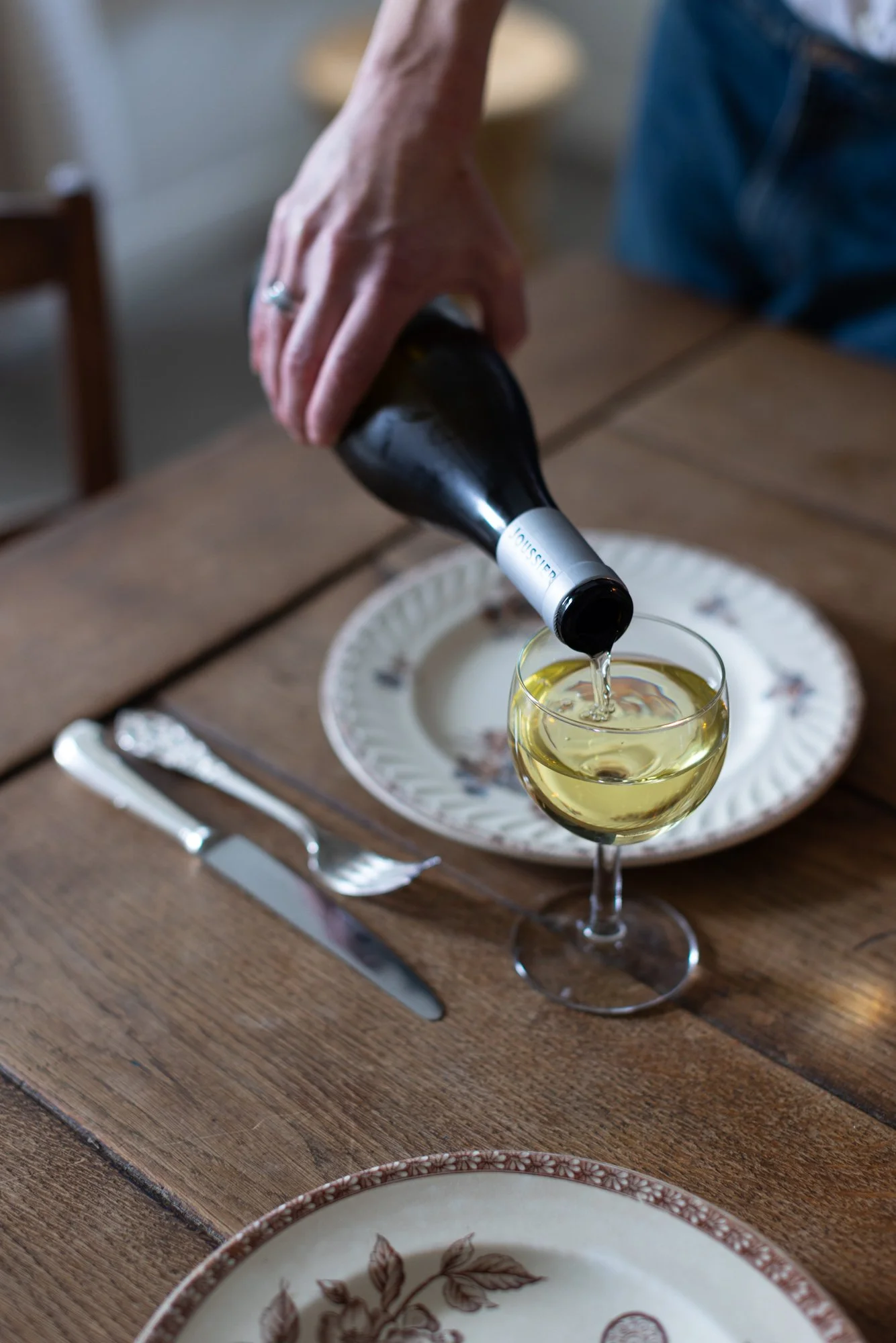 Person pouring white wine into a wine glass on a wooden table set with plates and cutlery.