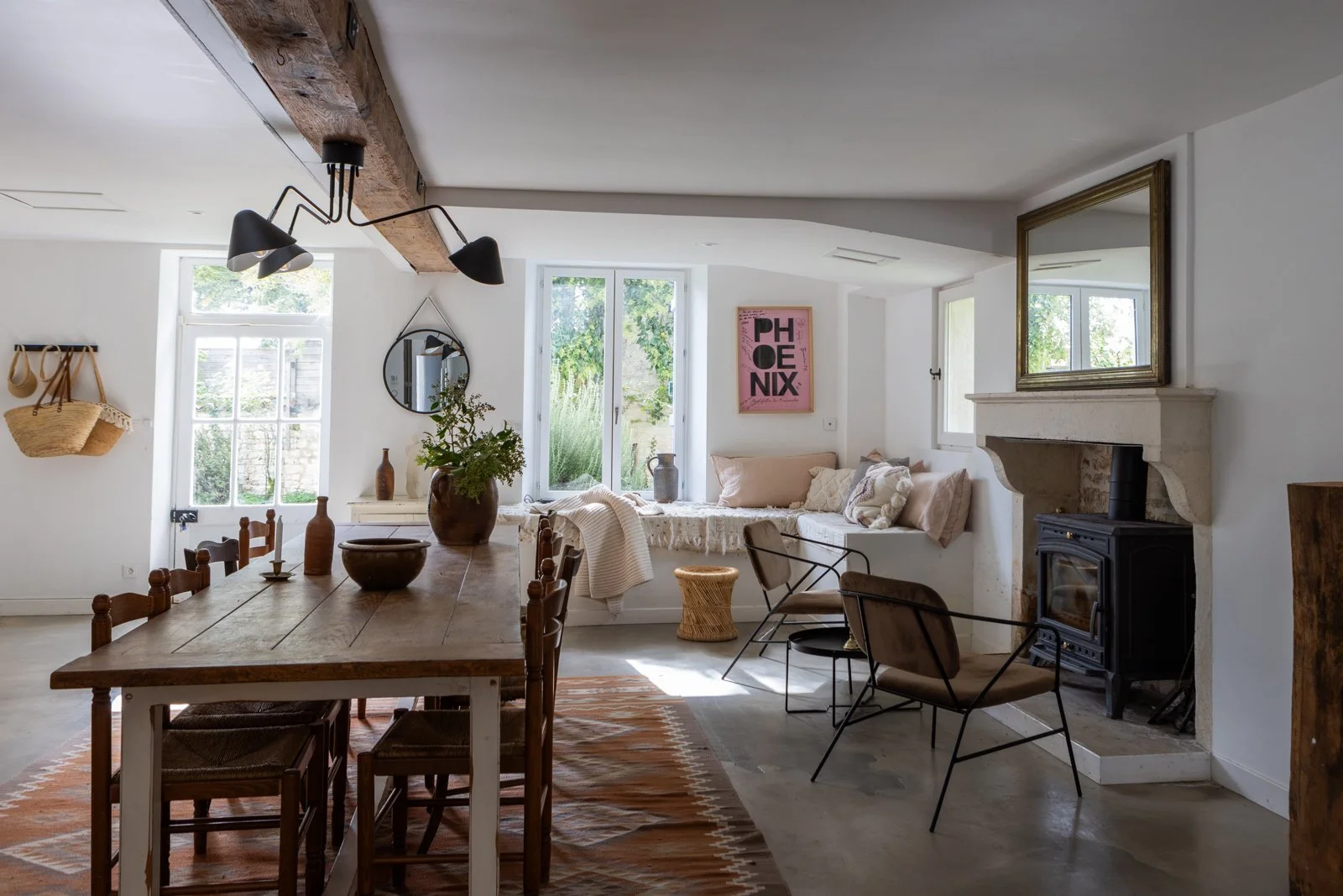 Bright living room with wooden dining table, metal chairs, fireplace, window seat with cushions, large mirror above fireplace, decorated with vases and plants, pink Phoenix poster on wall.