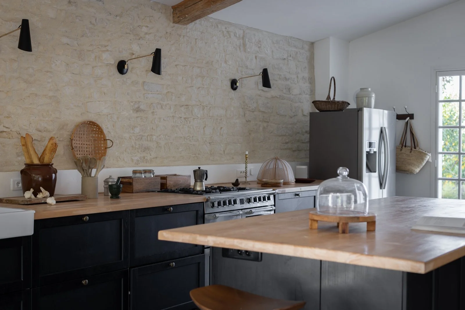 Kitchen with stone wall, black cabinets, wooden countertops, a stainless steel stove, a refrigerator, various kitchen items, and a window with natural light.