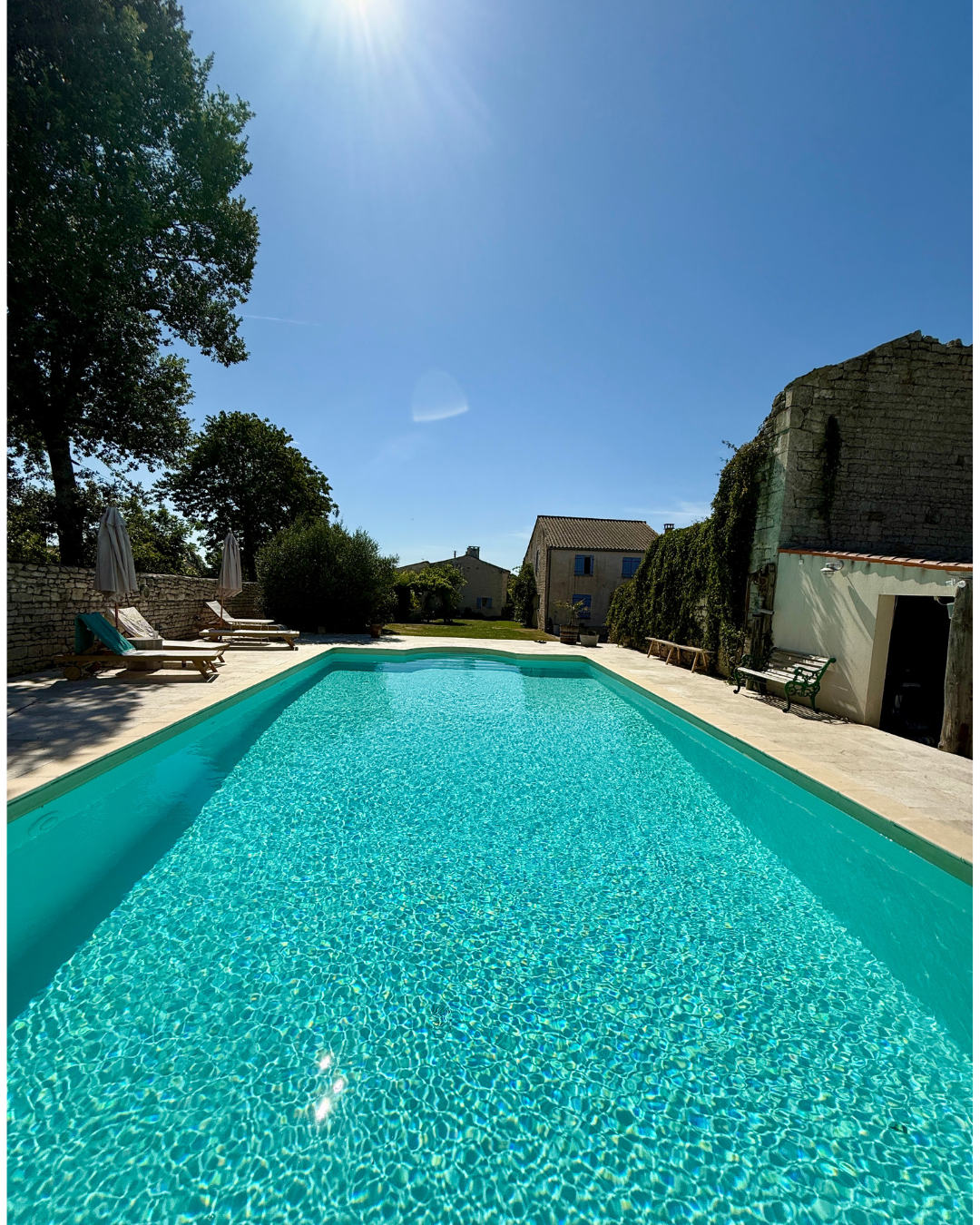 Sunny outdoor swimming pool with lounge chairs and umbrellas around it, clear blue water, stone wall, trees, and small buildings in background under a bright sky.