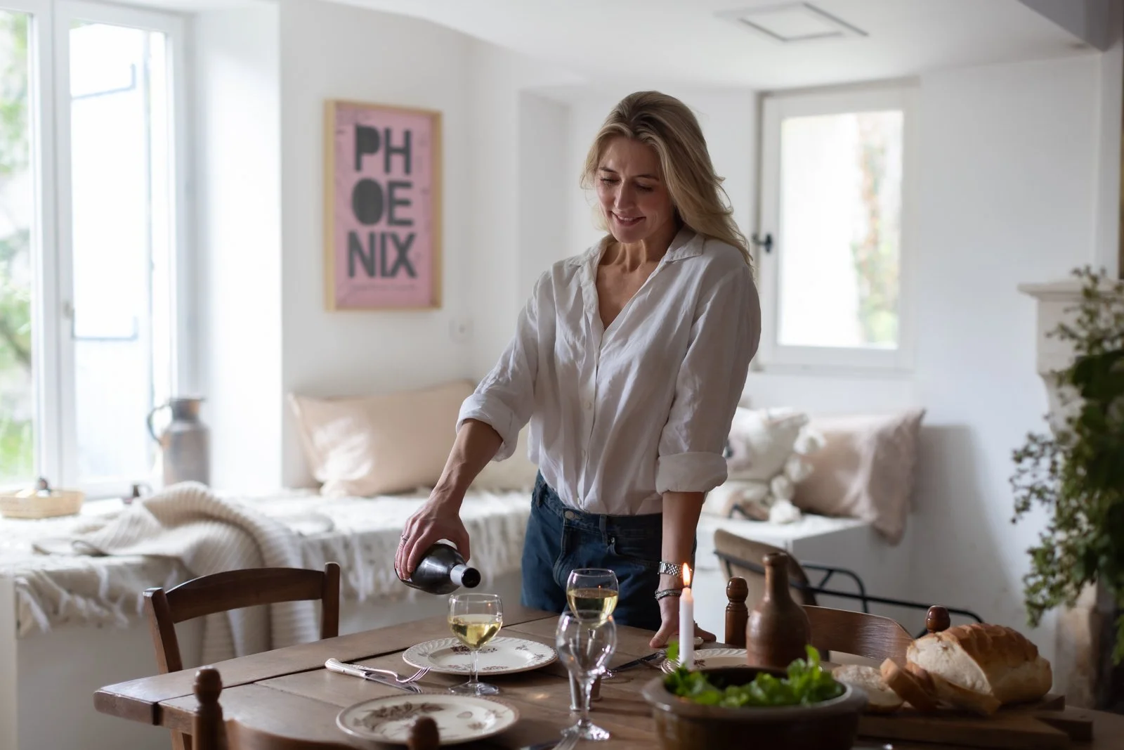 A woman in a white shirt pouring white wine into a glass at a dining table set for dinner in a bright, cozy home.