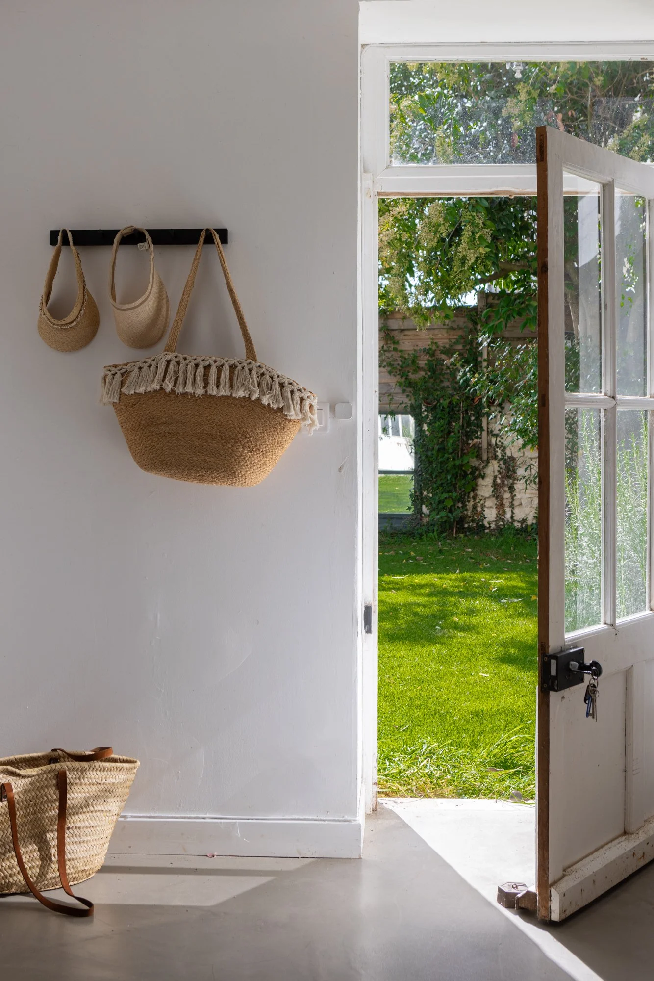 Open front door opens to a green lawn with trees and a garden wall outside, sunlight streaming in, inside a white-walled room with hooks holding straw bags hanging on the wall, and a woven straw tote bag on the floor.