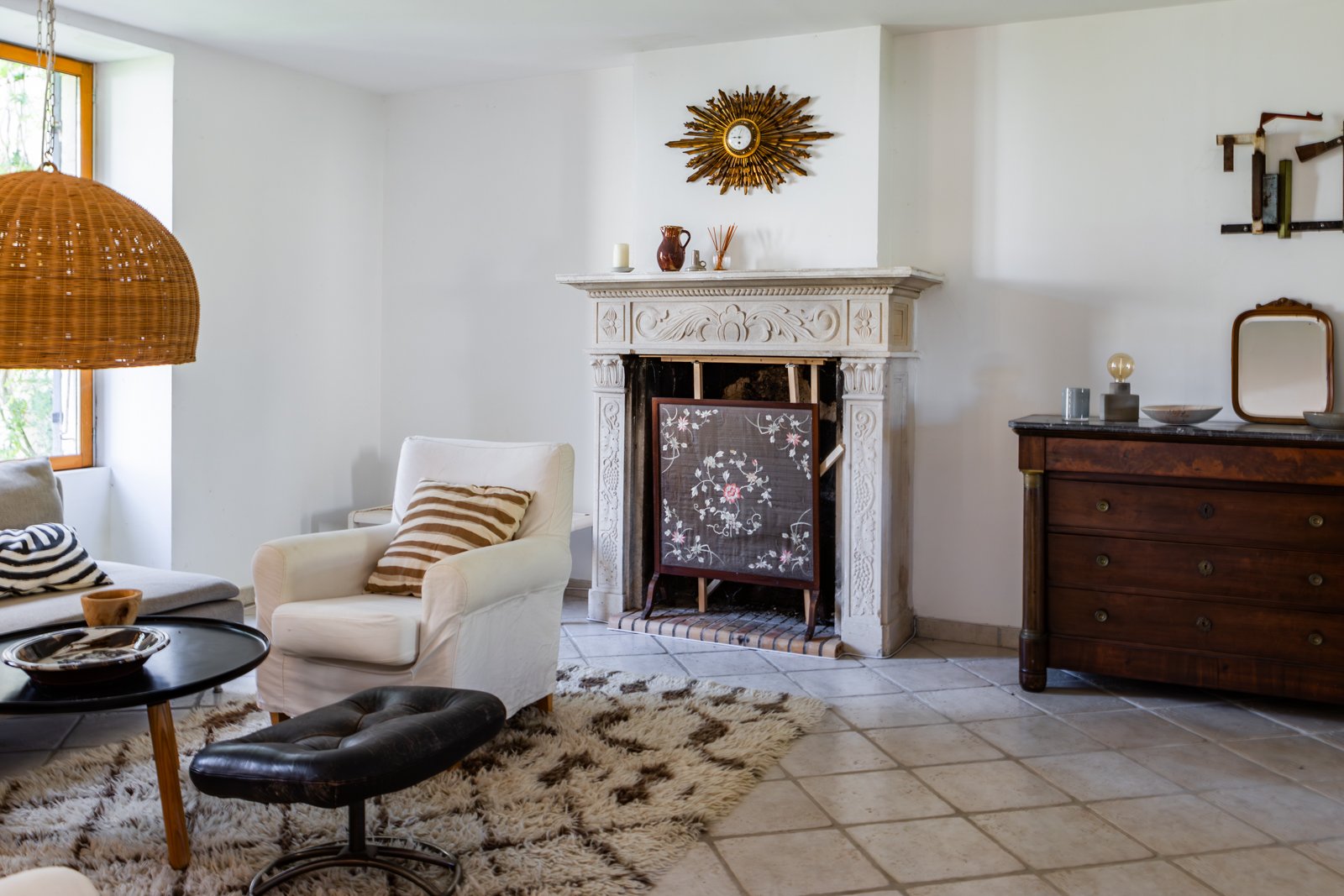 Living room with white walls, a fireplace, a white armchair with a striped cushion, a black coffee table with a bowl, a black ottoman, a dark wooden dresser with decorative items, a large window with a woven pendant lamp, and a floral room divider.