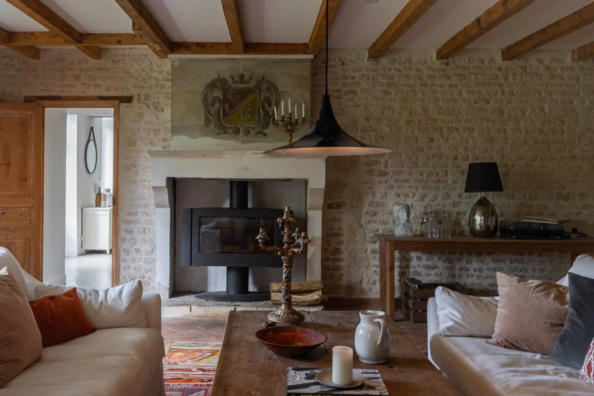 Living room with beige sofas, pillows, a wooden coffee table with a candelabra, candle, pitcher, and bowl. Stone walls and wood beams ceiling, a fireplace, and decorative items including a bust and a table lamp.