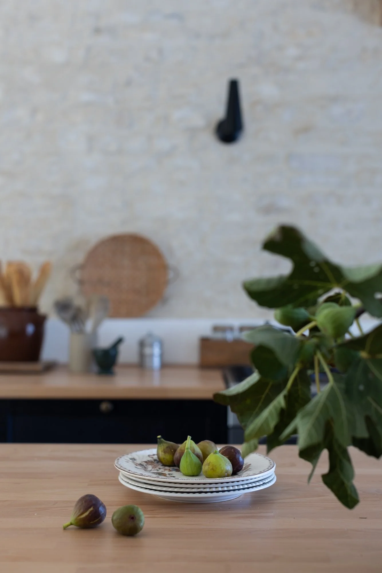 Figs on a decorative plate on a wooden kitchen counter with a bunch of green leaves to the right, kitchen utensils and a clock on the white brick wall in the background.