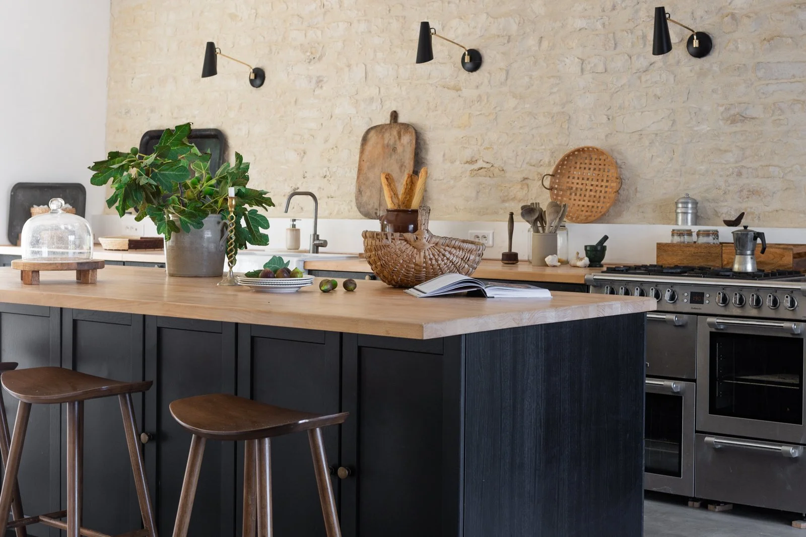 A modern kitchen with a large wooden island countertop, a potted plant, an open cookbook, baskets, and various kitchen utensils, set against a textured stone wall.
