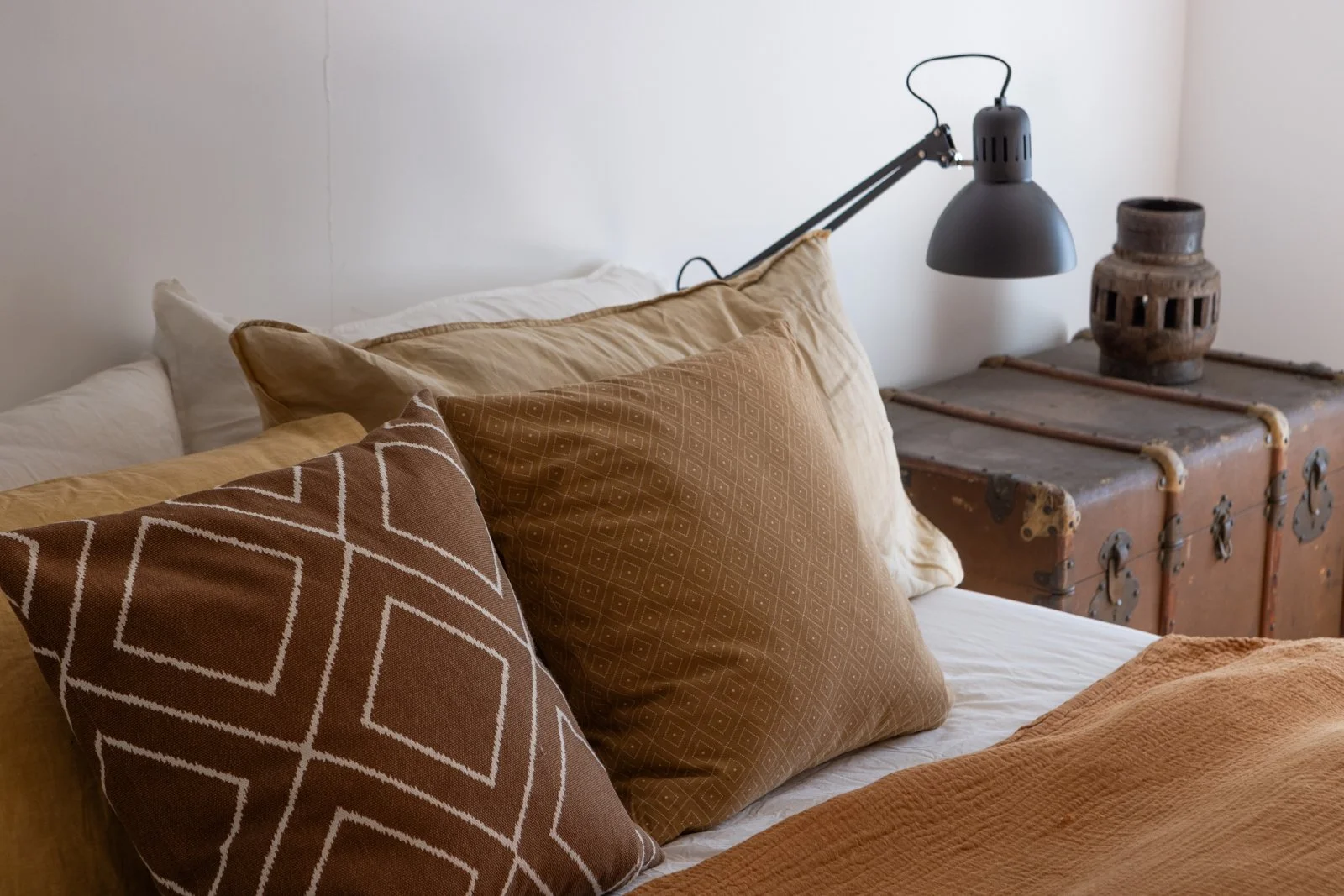 A cozy bed with multiple brown and beige pillows, a white bedsheet, and a beige blanket. A black adjustable bedside lamp is mounted on the wall next to a vintage wooden trunk used as a nightstand, with a decorative ceramic vase on top.