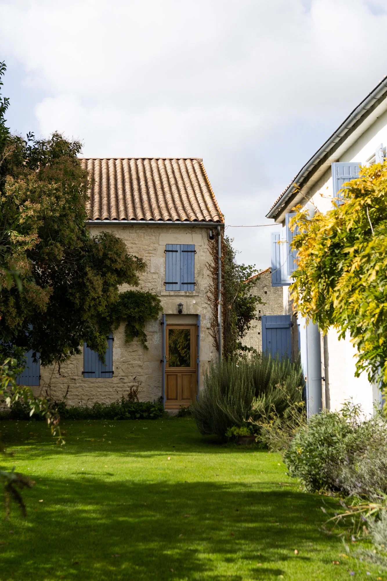A stone house with blue window shutters and a wooden door, surrounded by greenery and a well-maintained lawn in a garden.