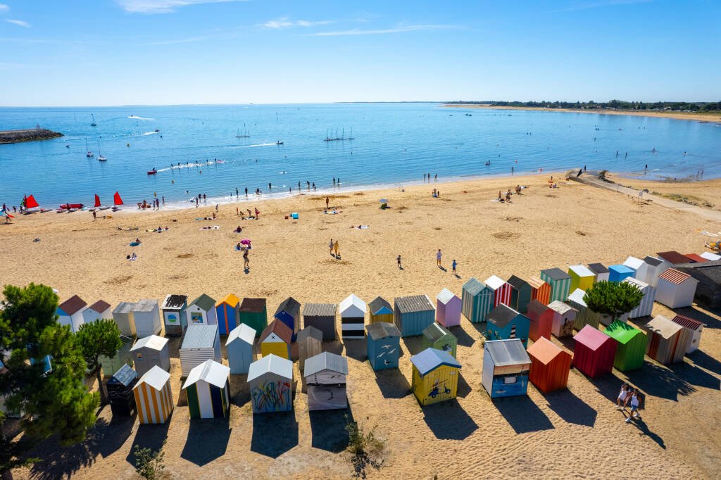 Beach with colorful beach huts lined up at the front, sandy shore with people relaxing and walking, blue ocean with sailboats and small boats, clear sky, and distant shoreline.
