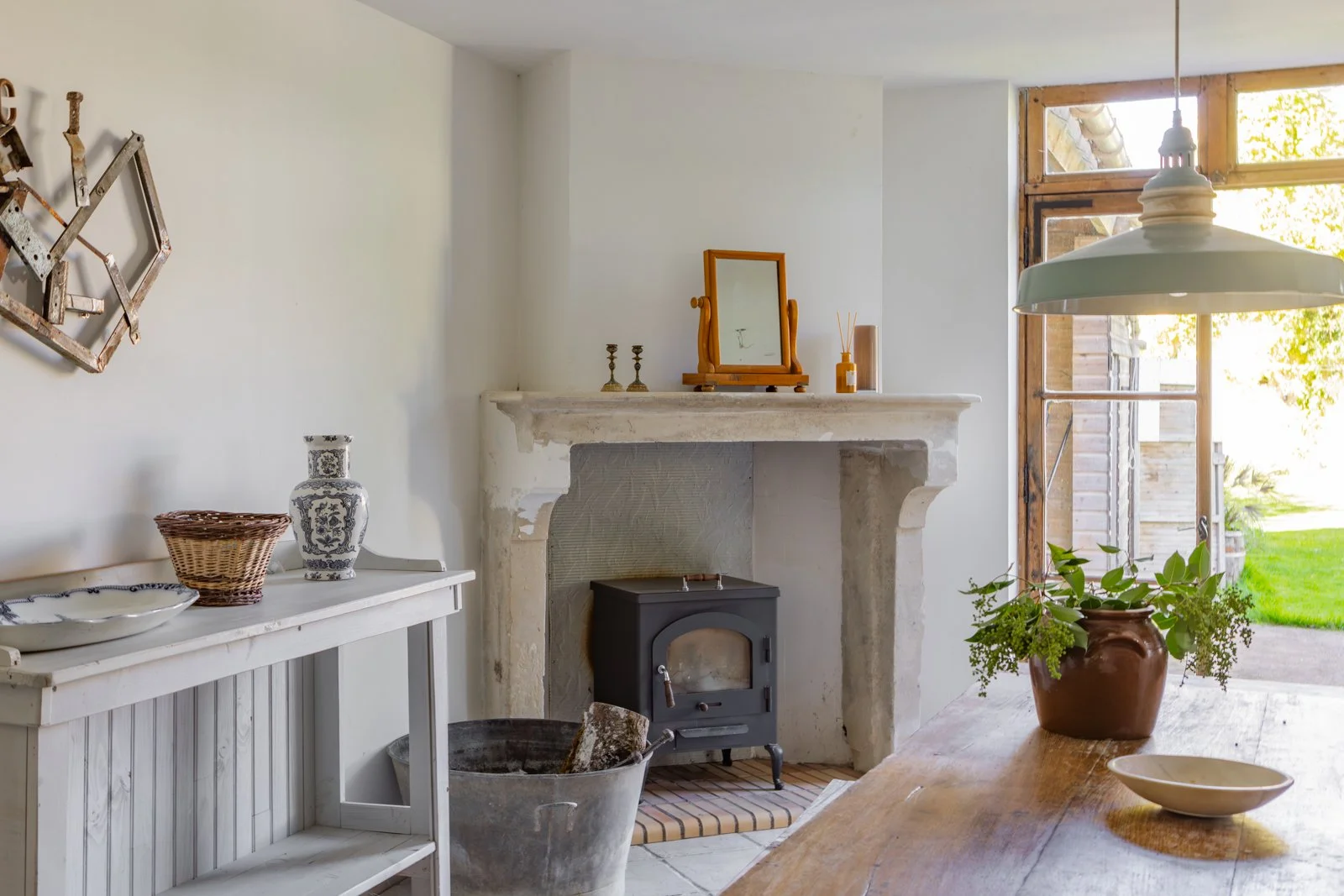 Cozy living room with a rustic fireplace, wooden table with a potted plant, and a view of the outdoors through large windows.