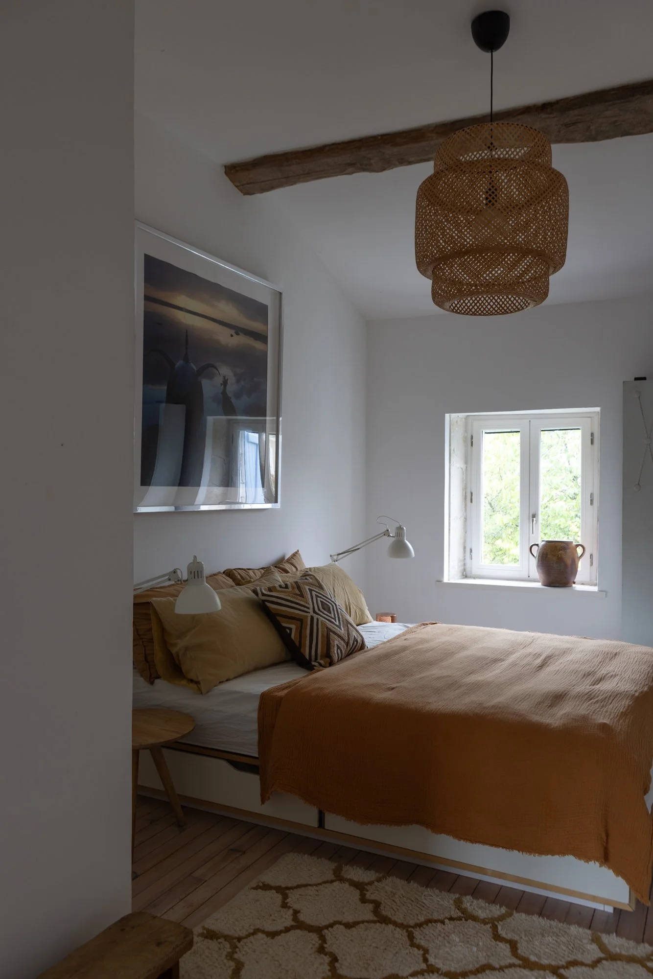A cozy bedroom with a double bed, beige bedding, and patterned pillows. A window with greenery outside, a potted plant on the window sill, a wooden side table, and a rattan ceiling lamp.